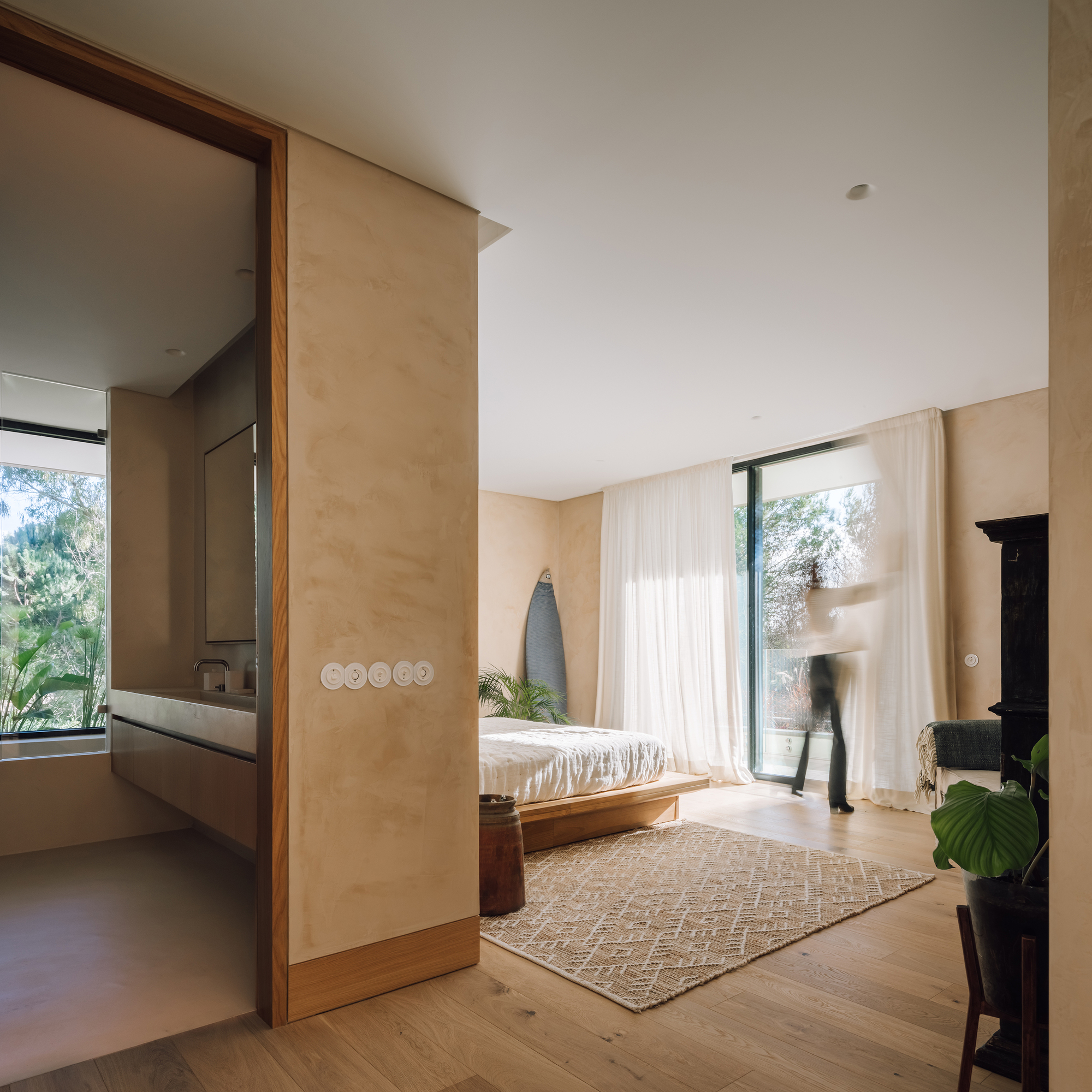 Bedroom suite with ensuite bathroom visible through timber-framed opening as figure passes glazed door