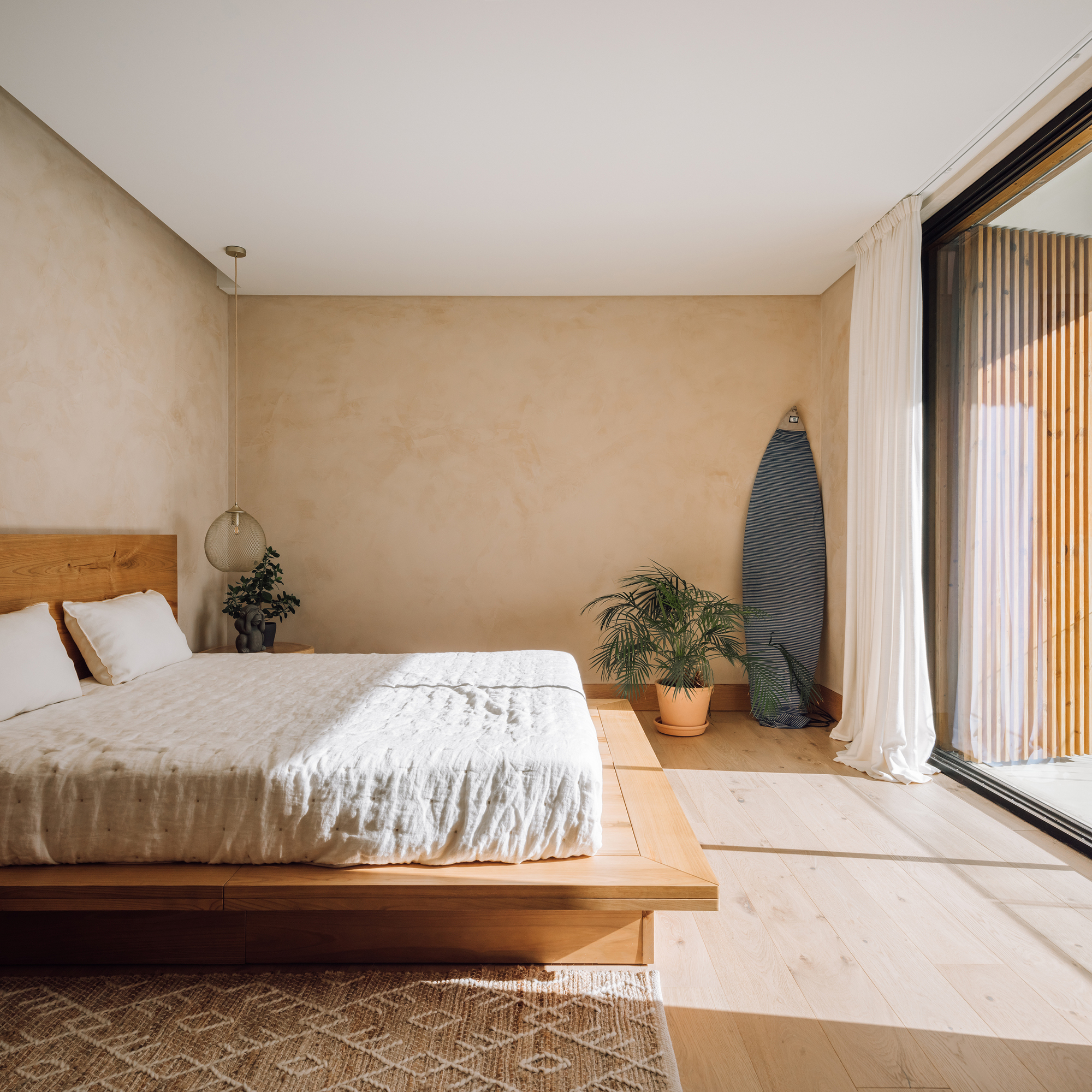 Platform bed in room with lime-washed walls and surfboard leaning beside floor-to-ceiling glazing