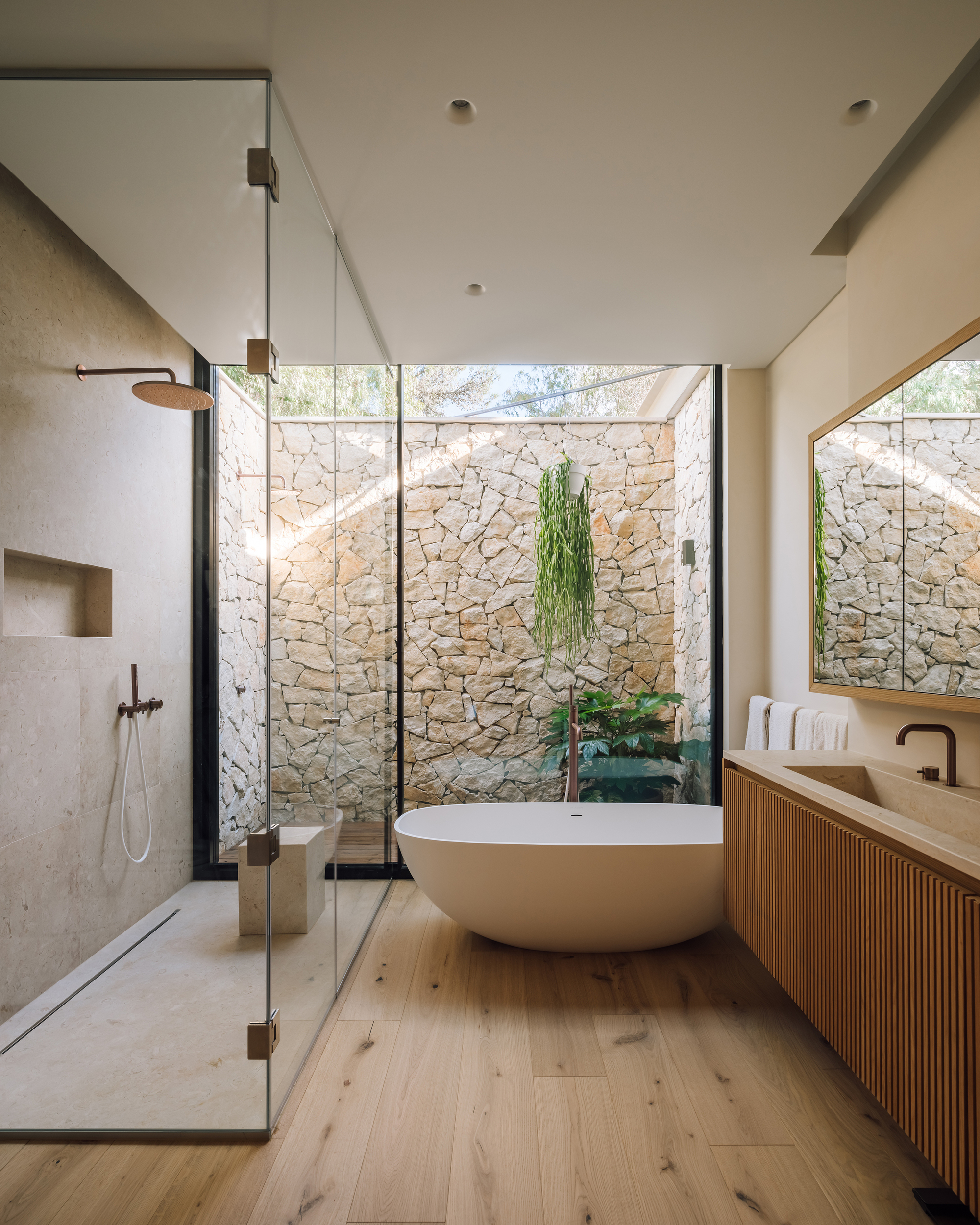 Bathroom with freestanding tub and glass-enclosed shower facing stone courtyard wall with hanging plants