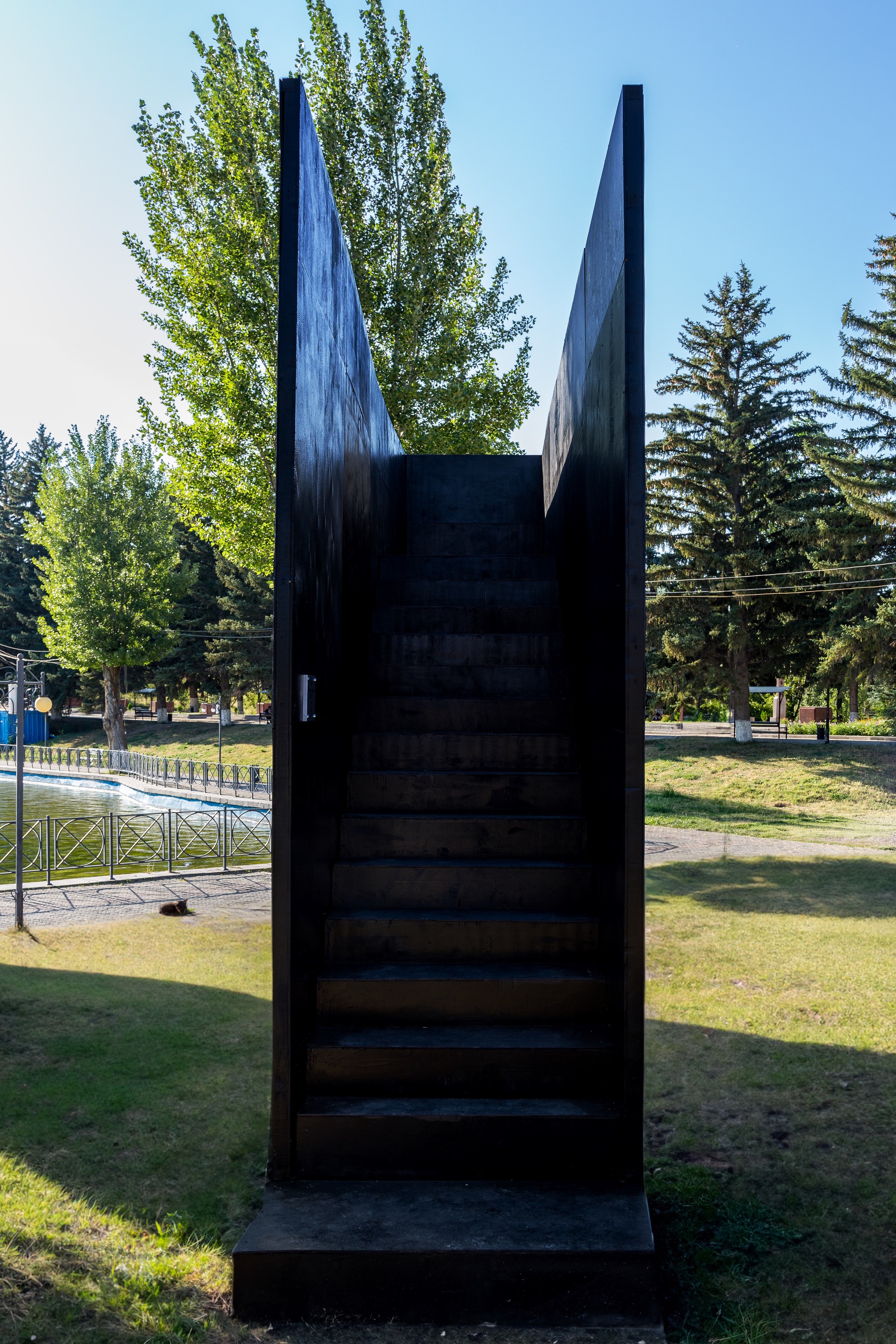 Dark metal staircase leading up between two angled mirrored panels surrounded by park landscape
