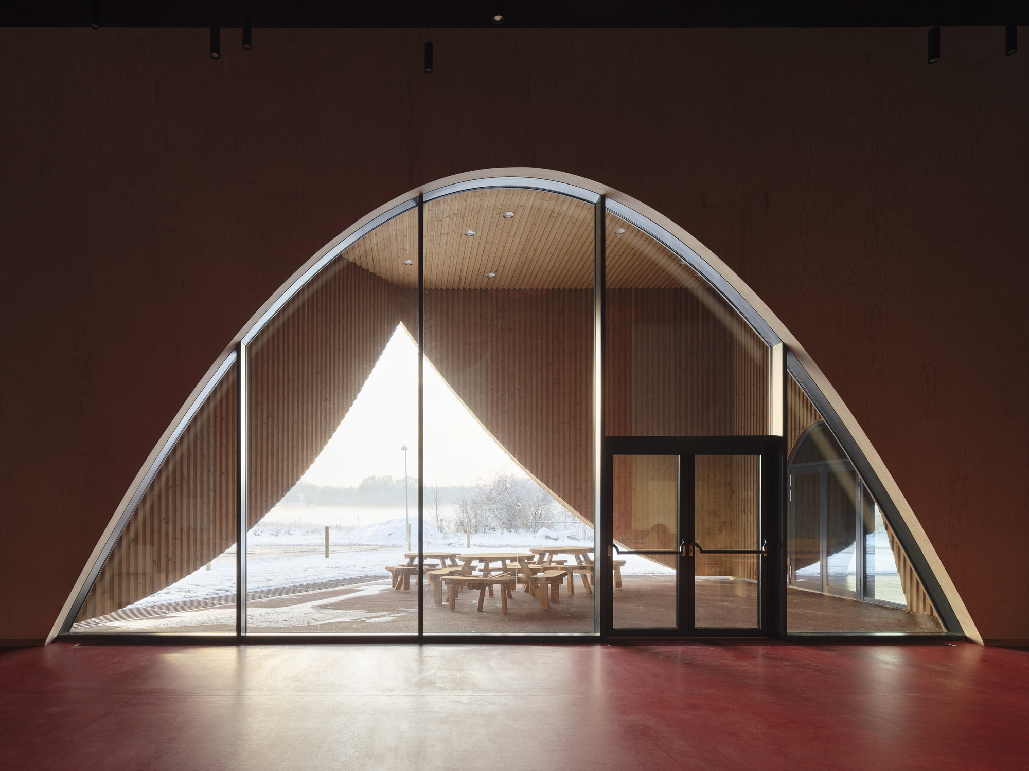 Arched glazed opening framing a wood-lined porch and snowy landscape beyond a red interior floor