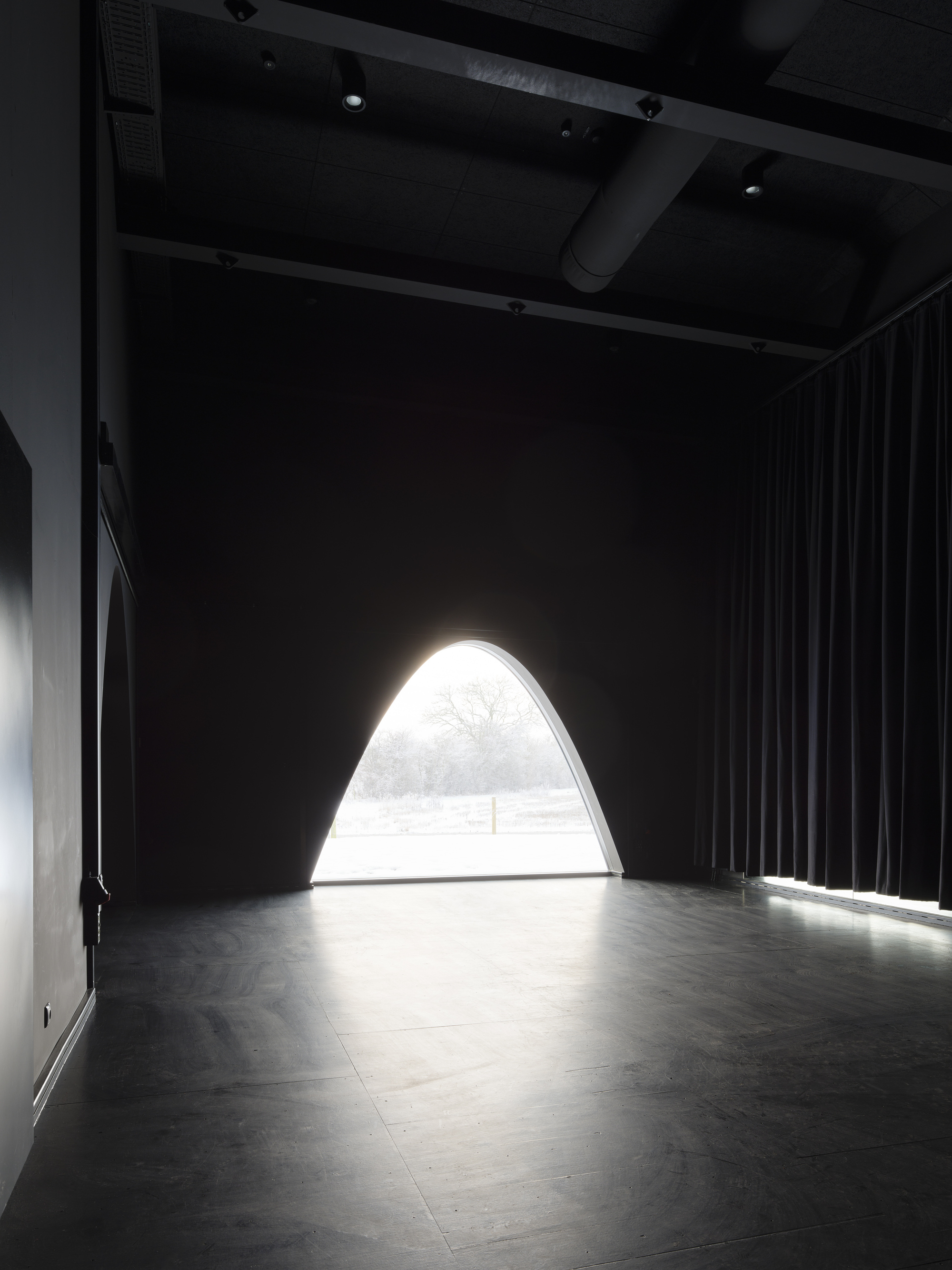 Arched opening flooding light into a dark performance space with black curtains and exposed ceiling
