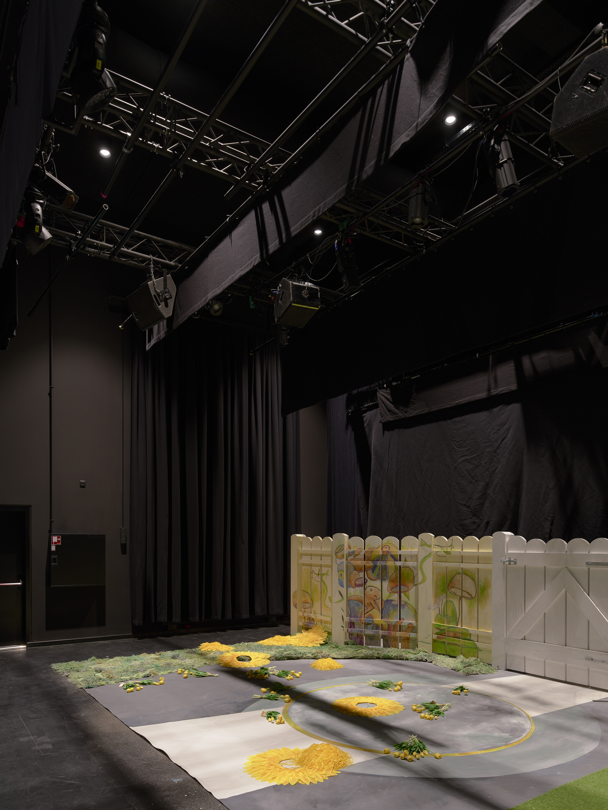 Black box theater space with exposed truss rigging and a stage floor painted with yellow and green patterns