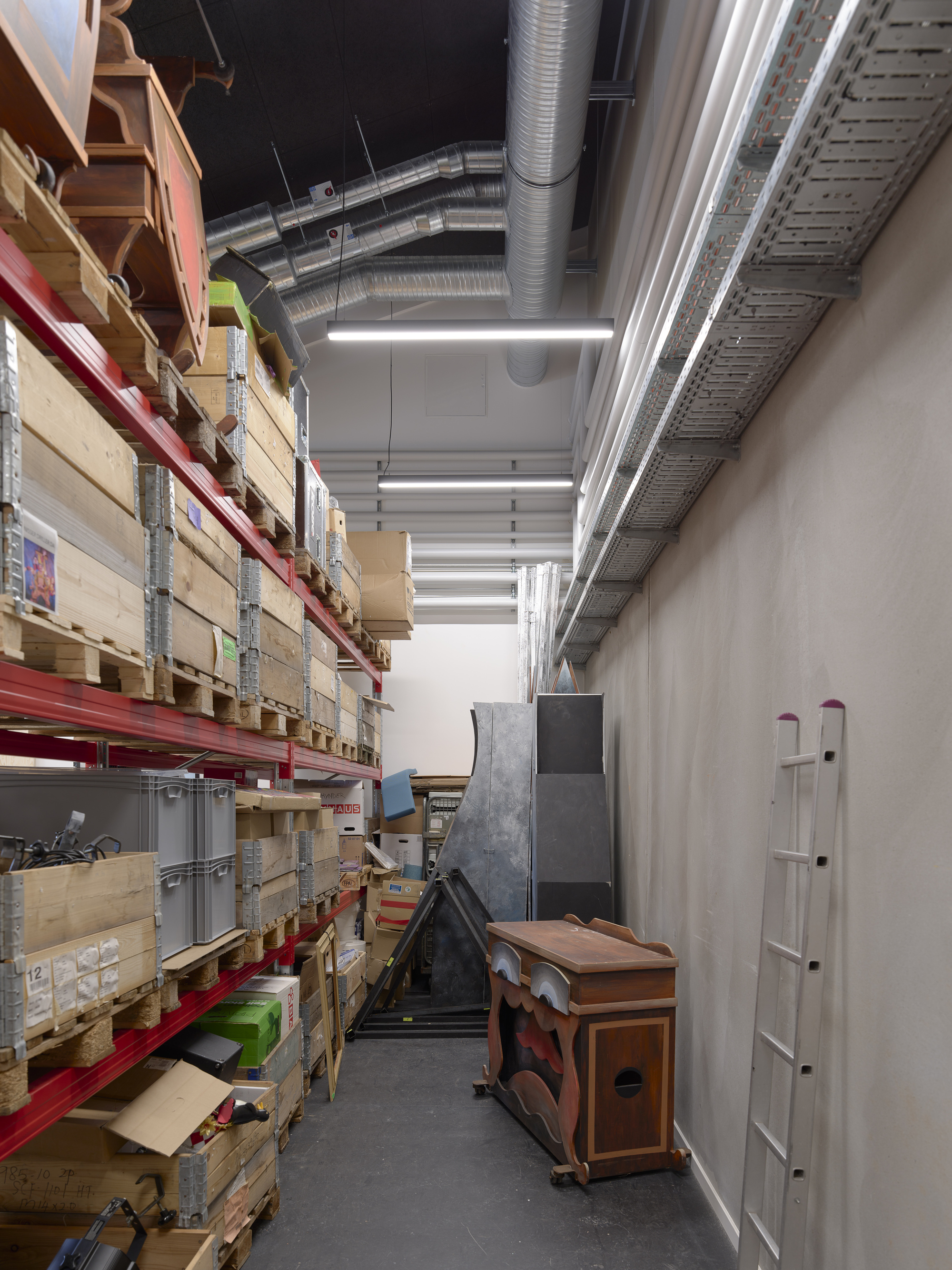 Storage corridor with red metal shelving units stacked with wooden crates below exposed ceiling ducts