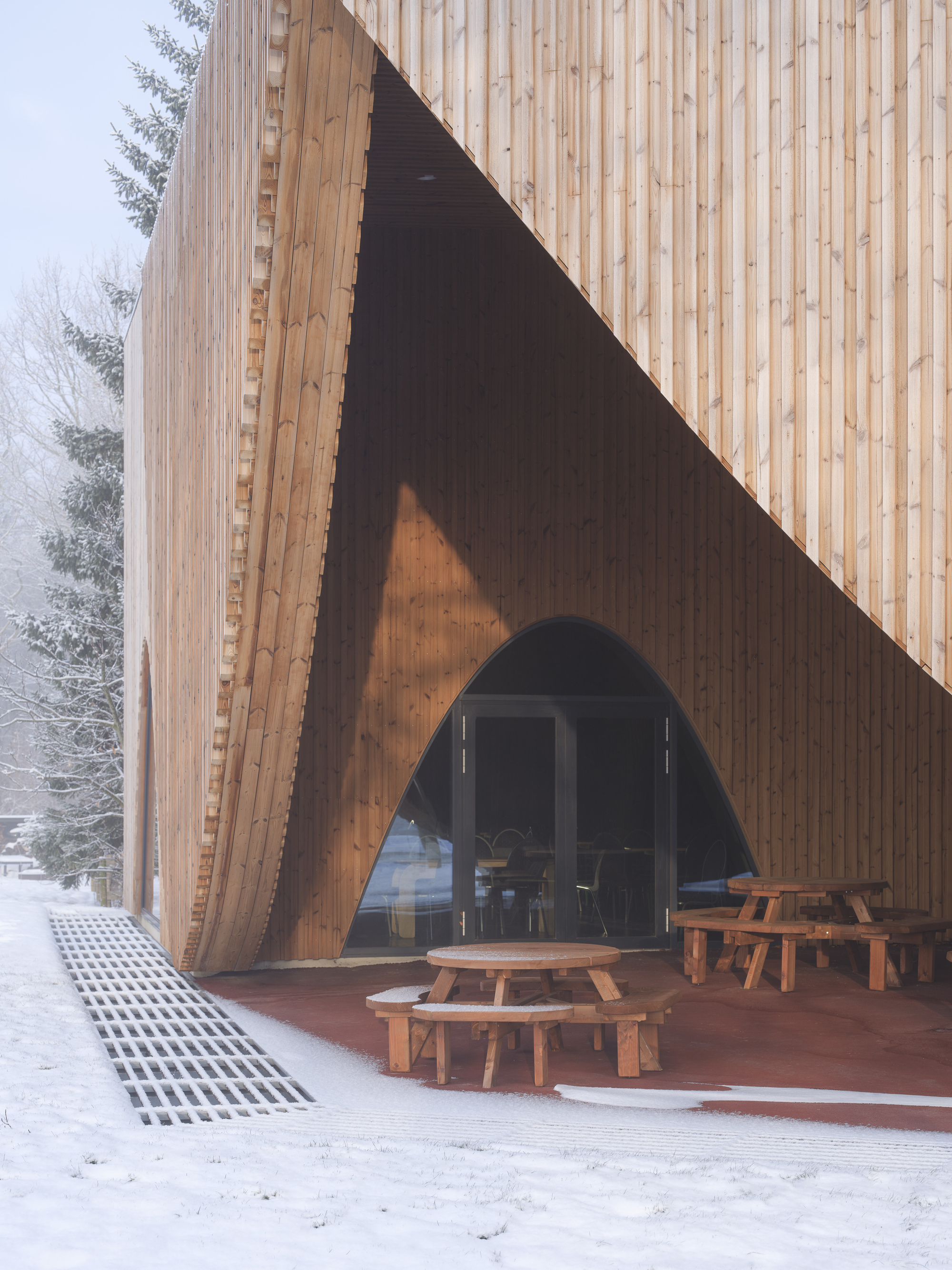 Triangular entrance opening framed by timber cladding with picnic benches on the covered terrace in snow