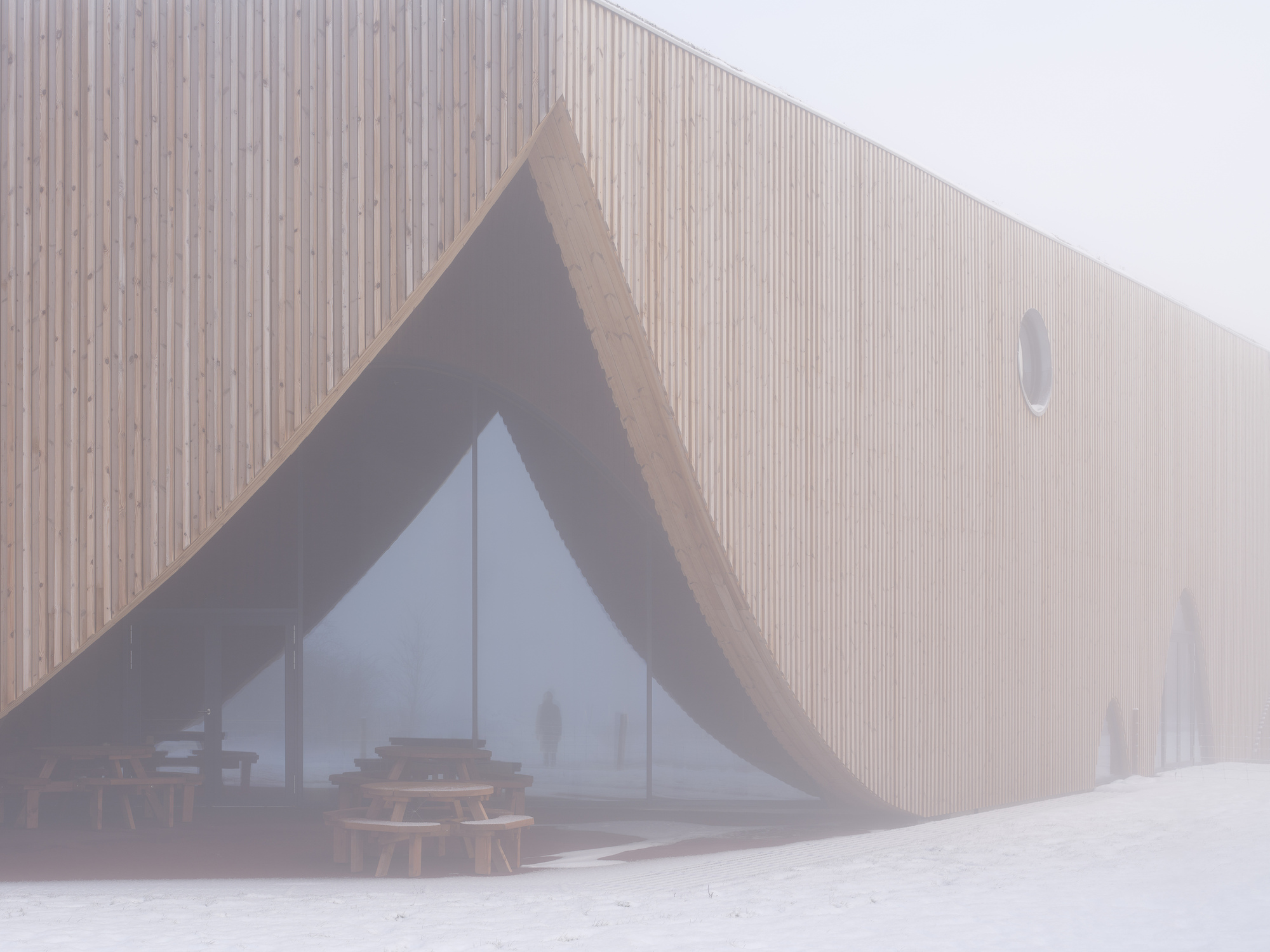 Timber-clad facade with triangular glazed entrance in heavy fog and fresh snow
