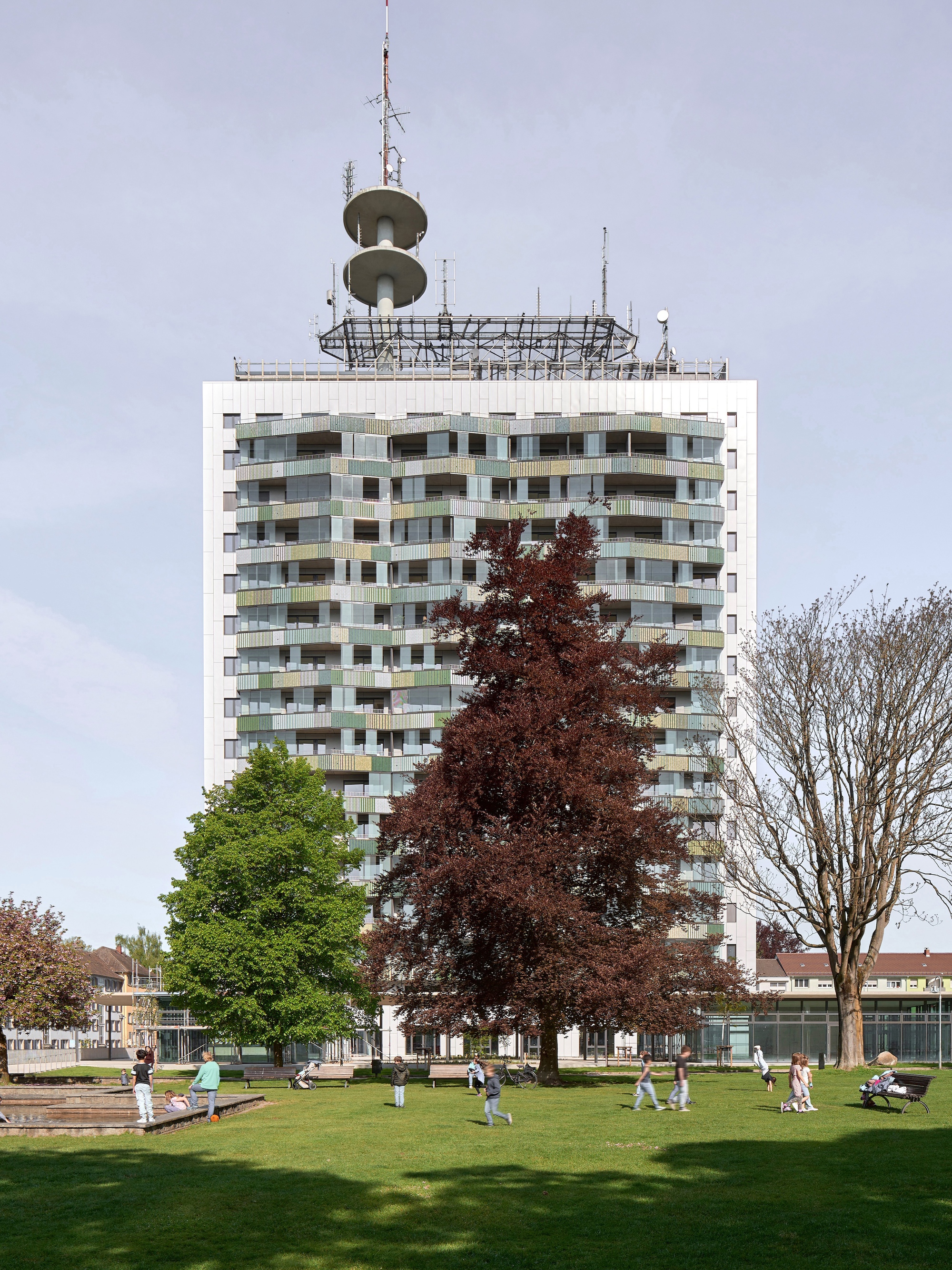 Tower with multicolored balcony grid rising above a public lawn with spring trees and scattered visitors