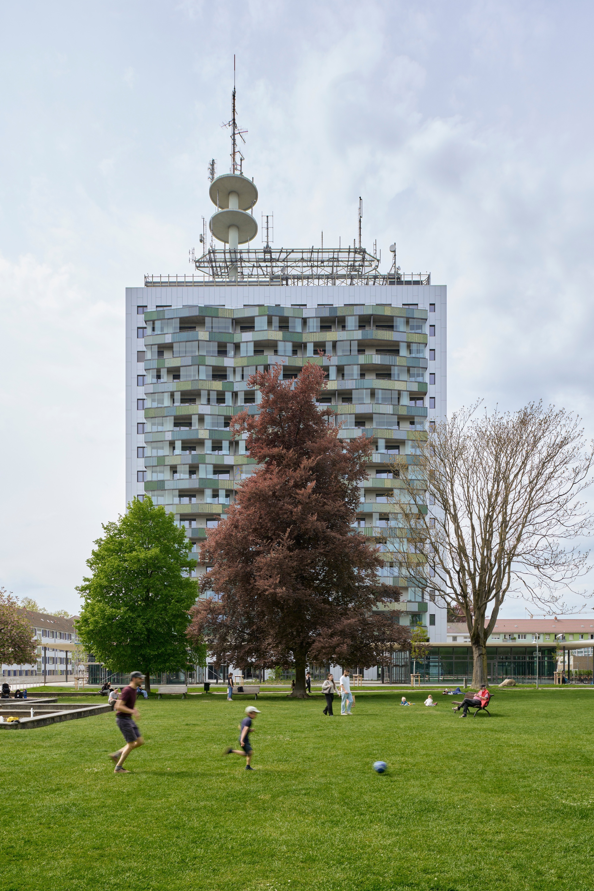 White tower with multicoloured balconies and rooftop antenna array behind a park lawn with children playing