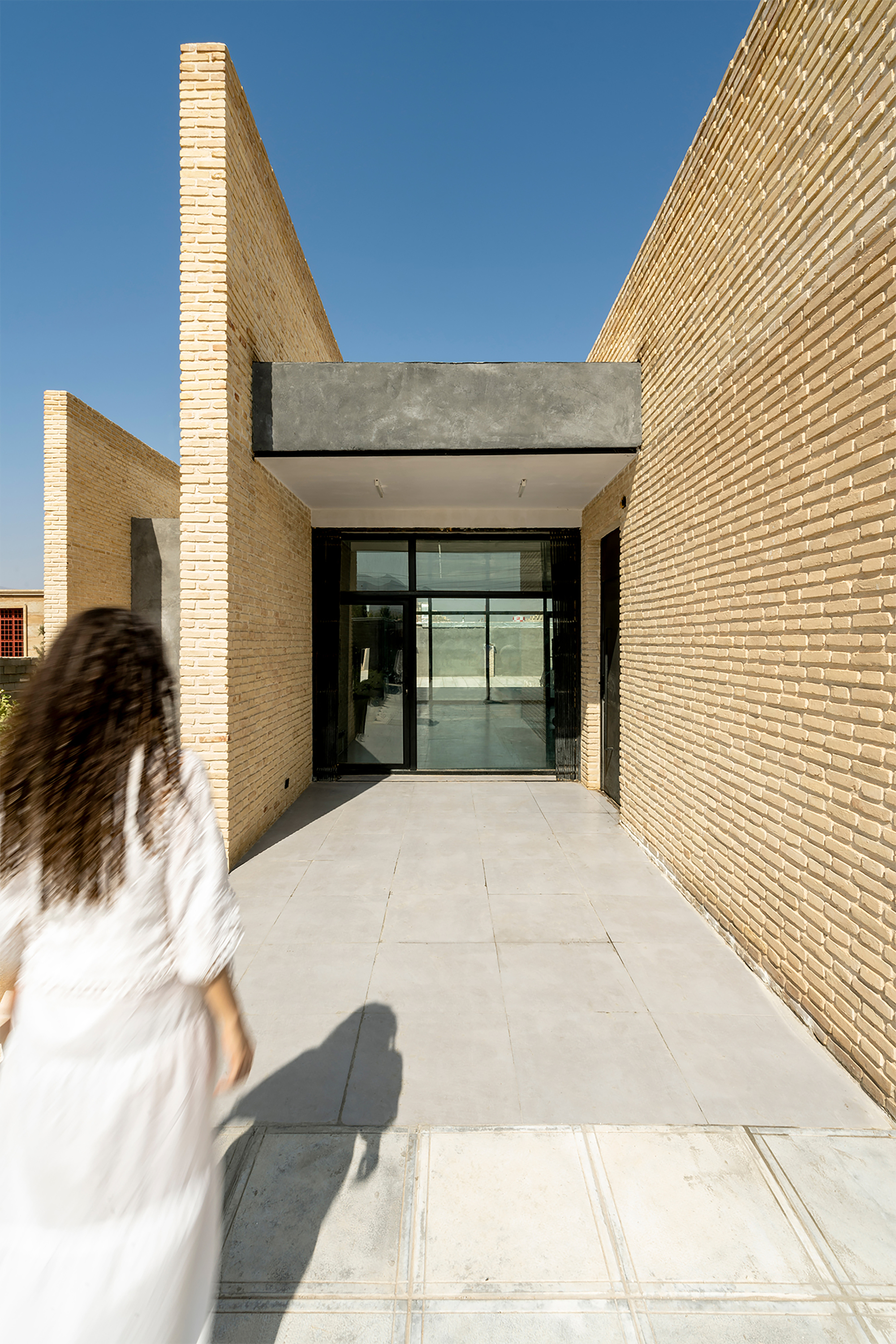 Entrance passageway lined with textured buff brick walls leading to glass doors under a concrete canopy