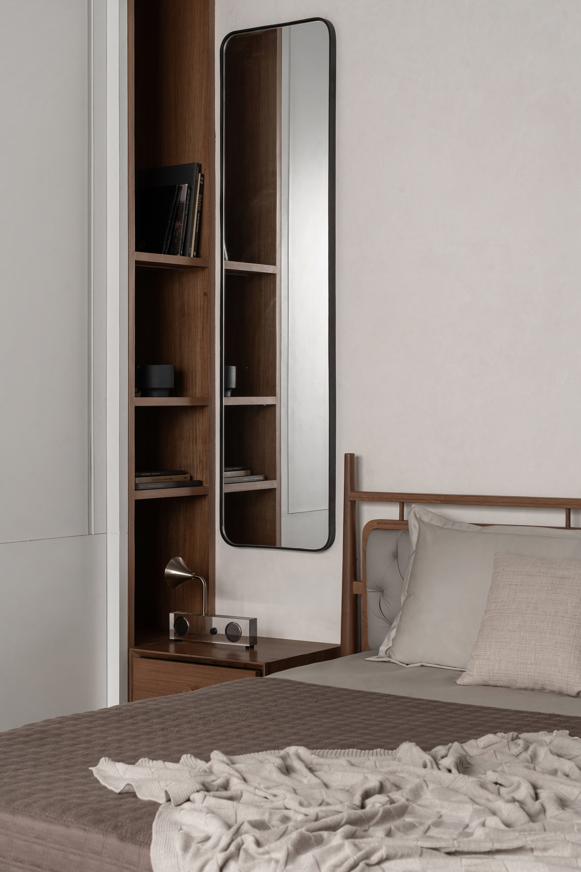 Tall standing mirror beside timber shelving niche and bed corner in soft daylight