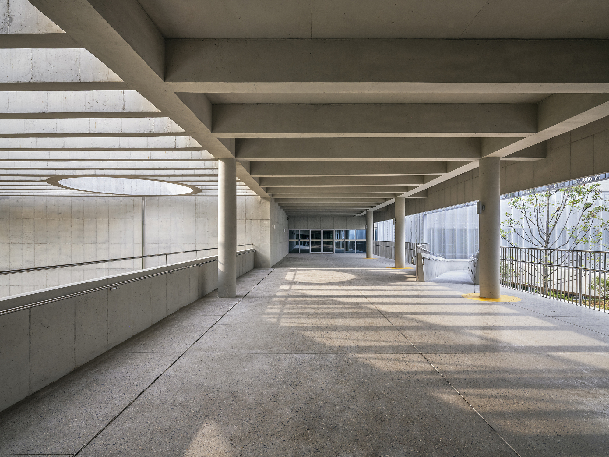Covered walkway with exposed concrete beam ceiling and dappled sunlight filtering through the lateral openings