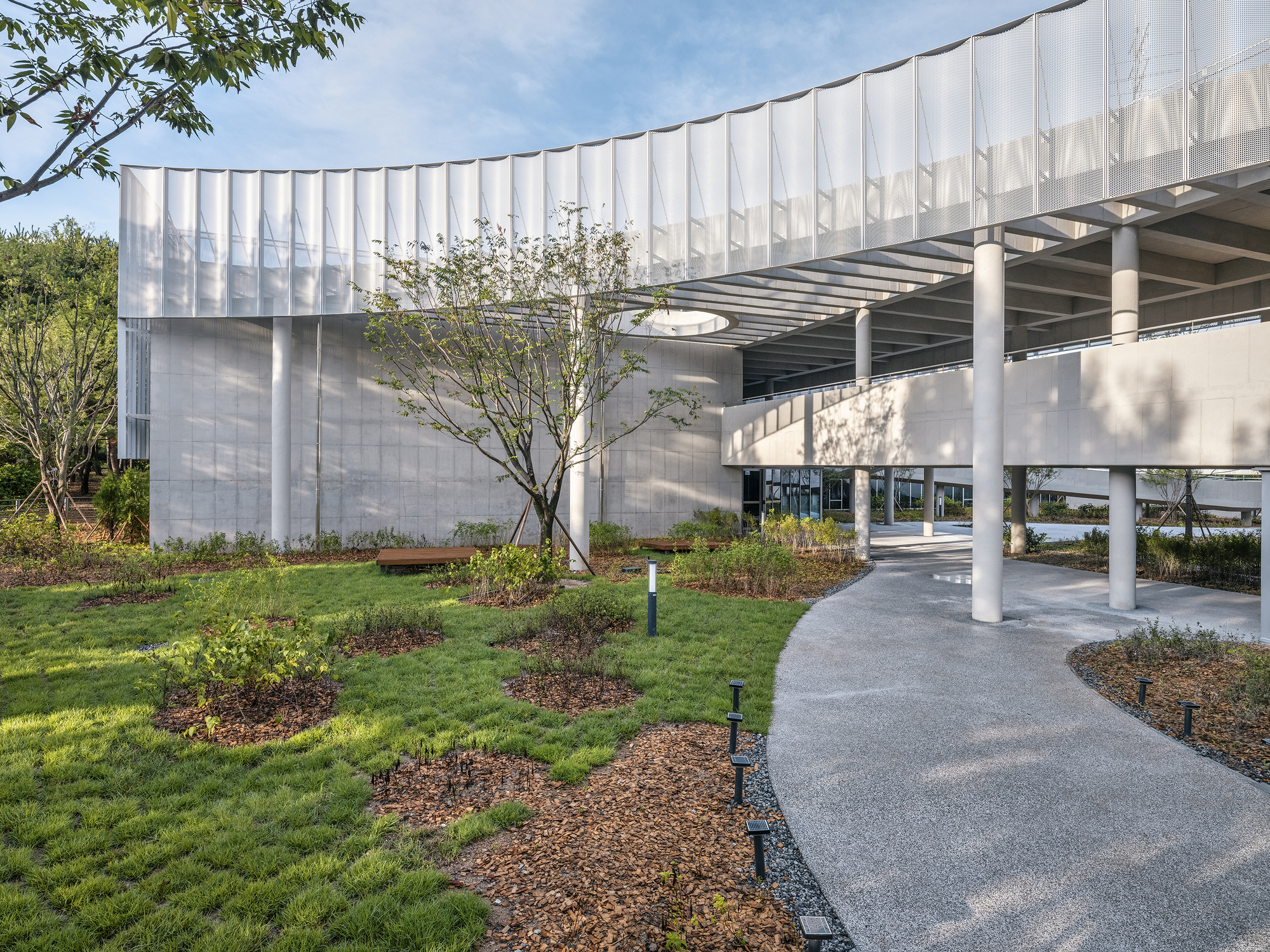 Covered walkway with white cylindrical columns beneath a raised platform and translucent facade panels