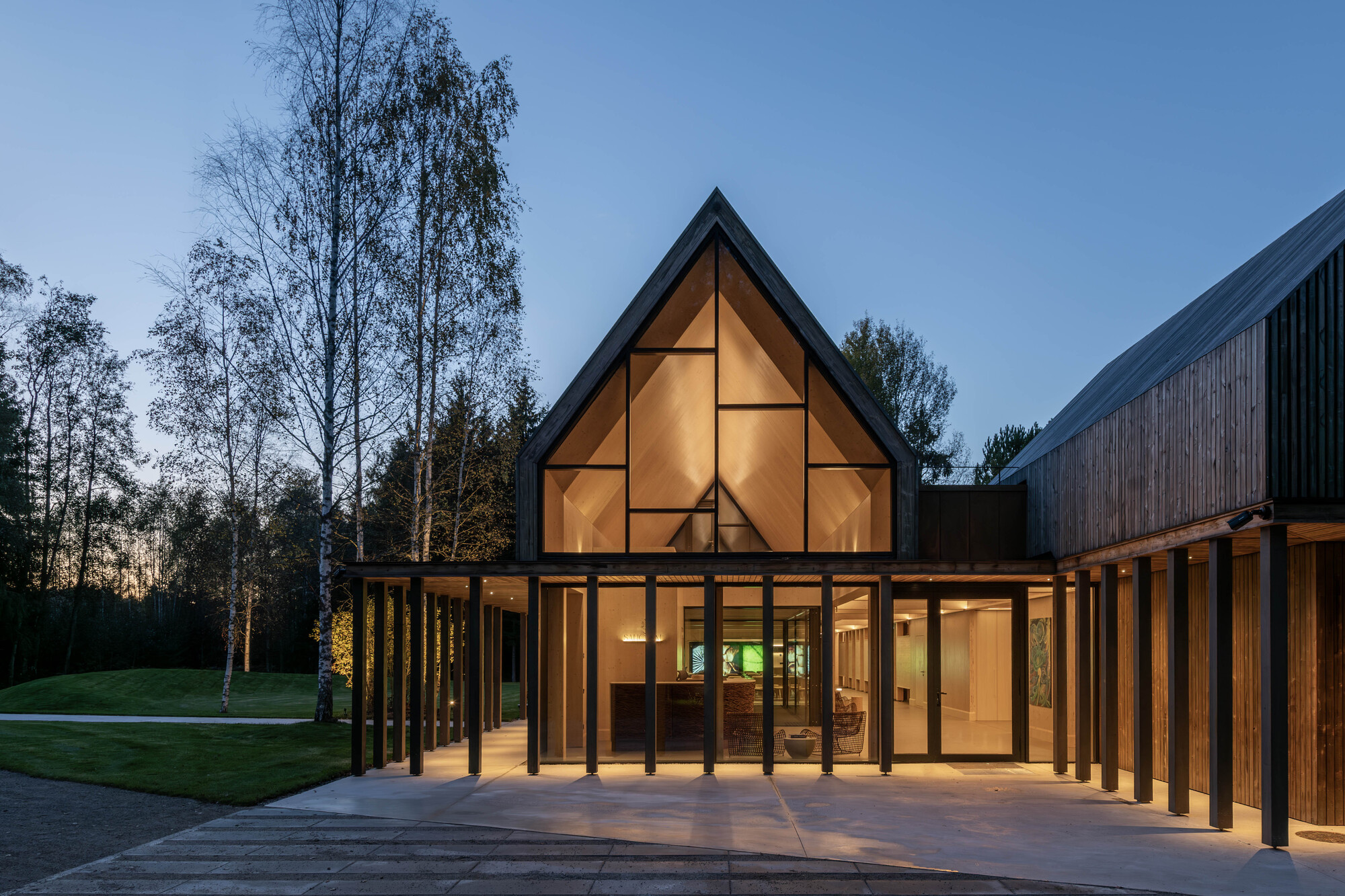 Covered terrace with timber columns connecting the glazed gable to adjacent wings at twilight