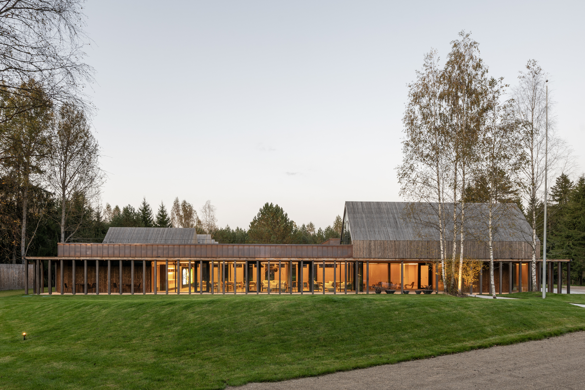 Long timber-clad volume with glazed openings and pitched roof volumes set in a lawn at dusk