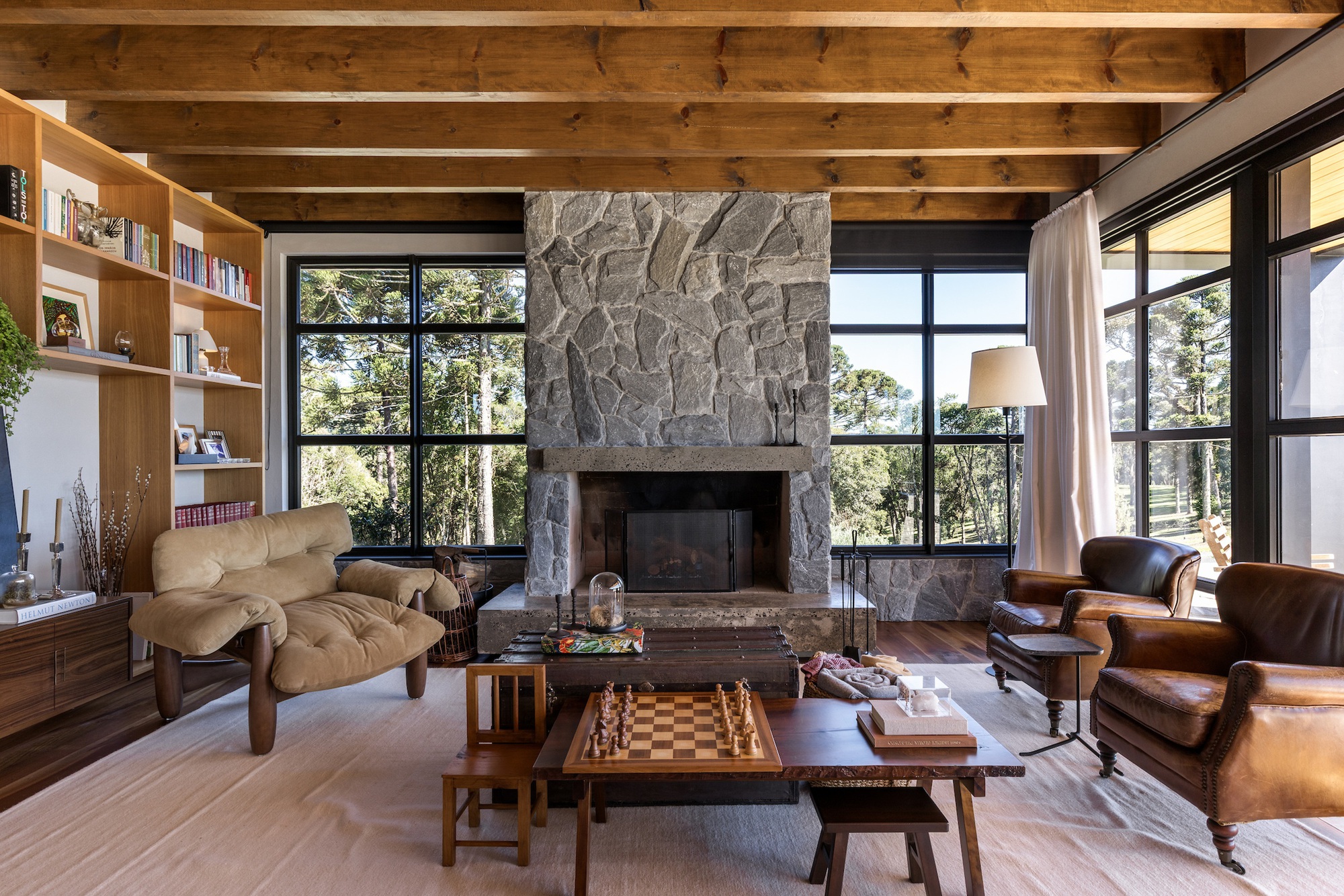 Living room with exposed timber ceiling beams, grey stone fireplace and black-framed windows overlooking trees