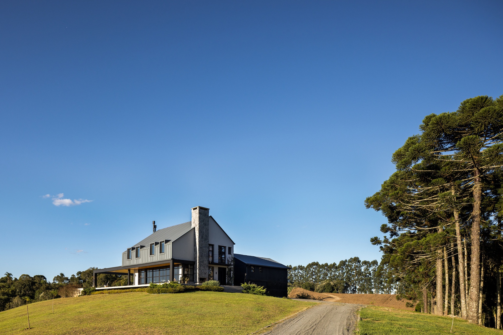 Two-story house with metal chimney and gravel driveway ascending a grassy slope near pines