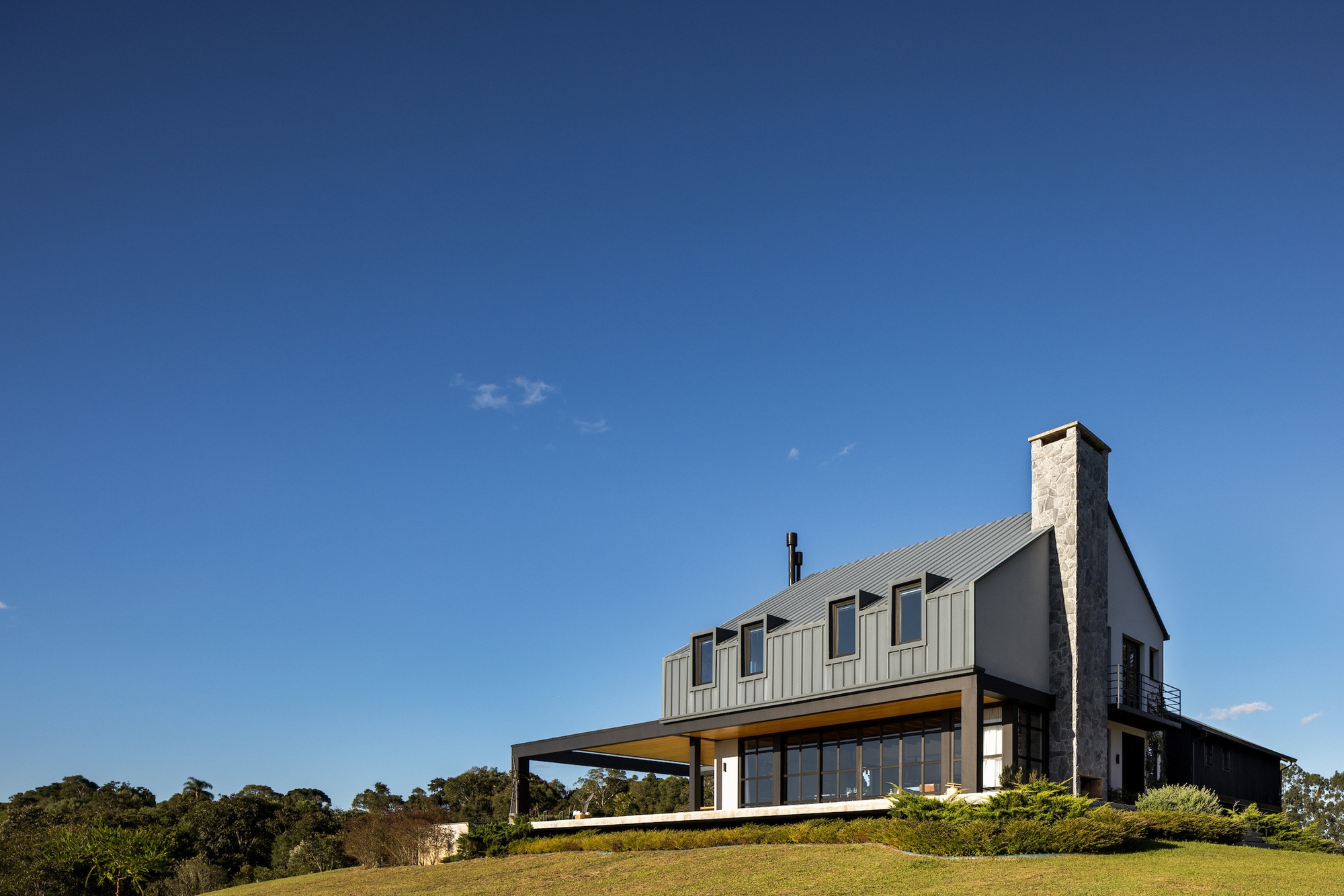 Two-storey gabled house with stone chimney and covered terrace set on a grassy hillside under clear blue sky