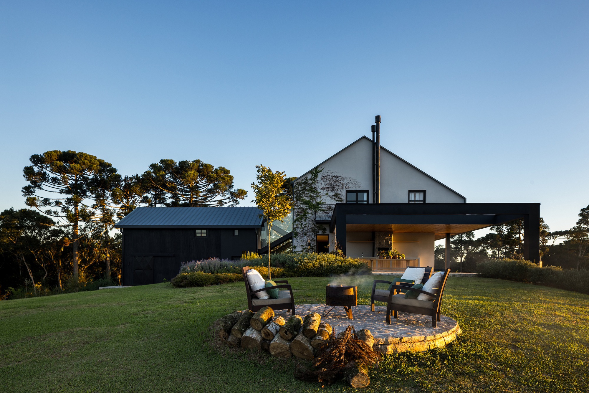 Stone fire pit with chairs on lawn facing the gabled house and outbuilding at dusk