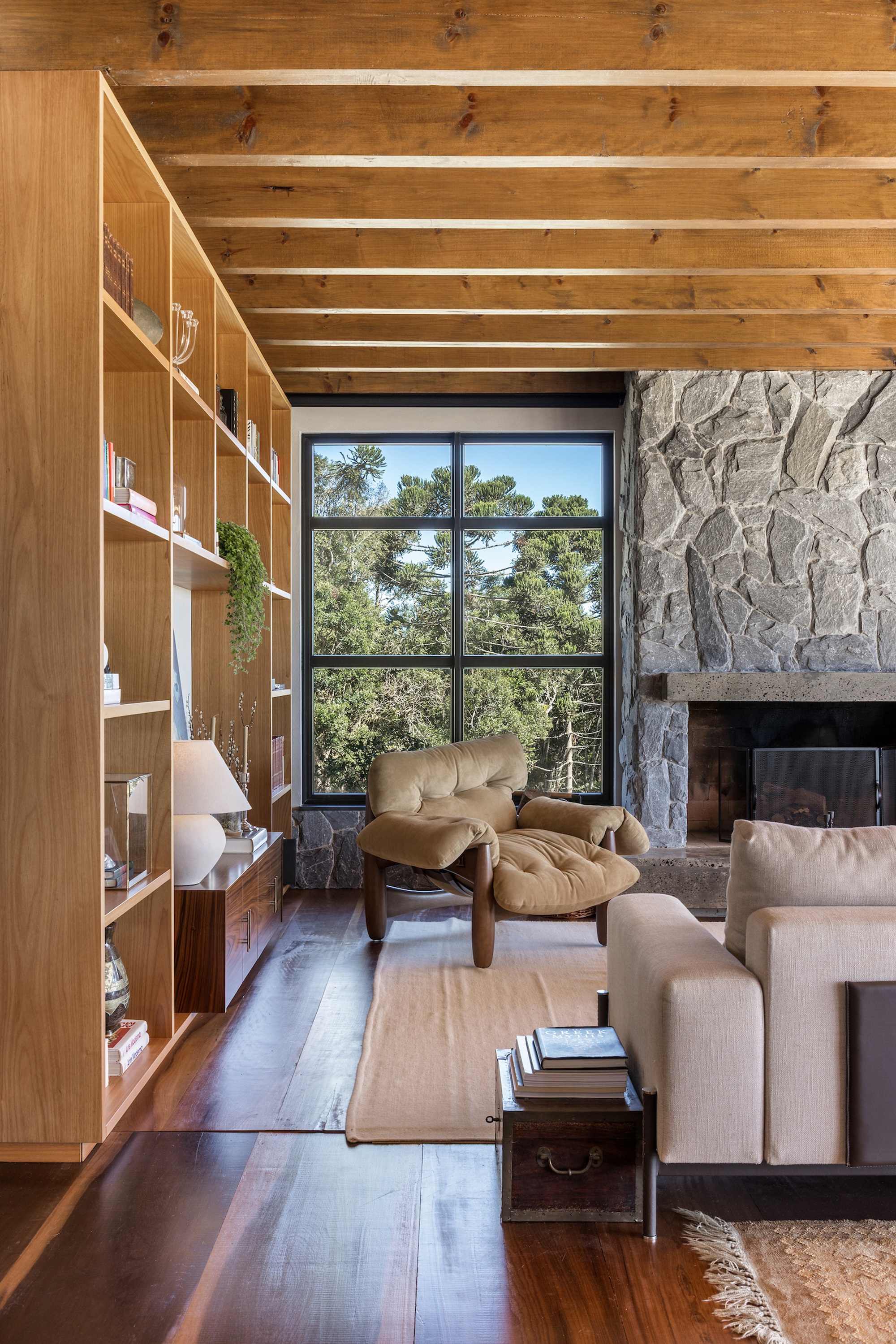 Living room with exposed timber beams, built-in shelving, and stone fireplace framing a view of treetops