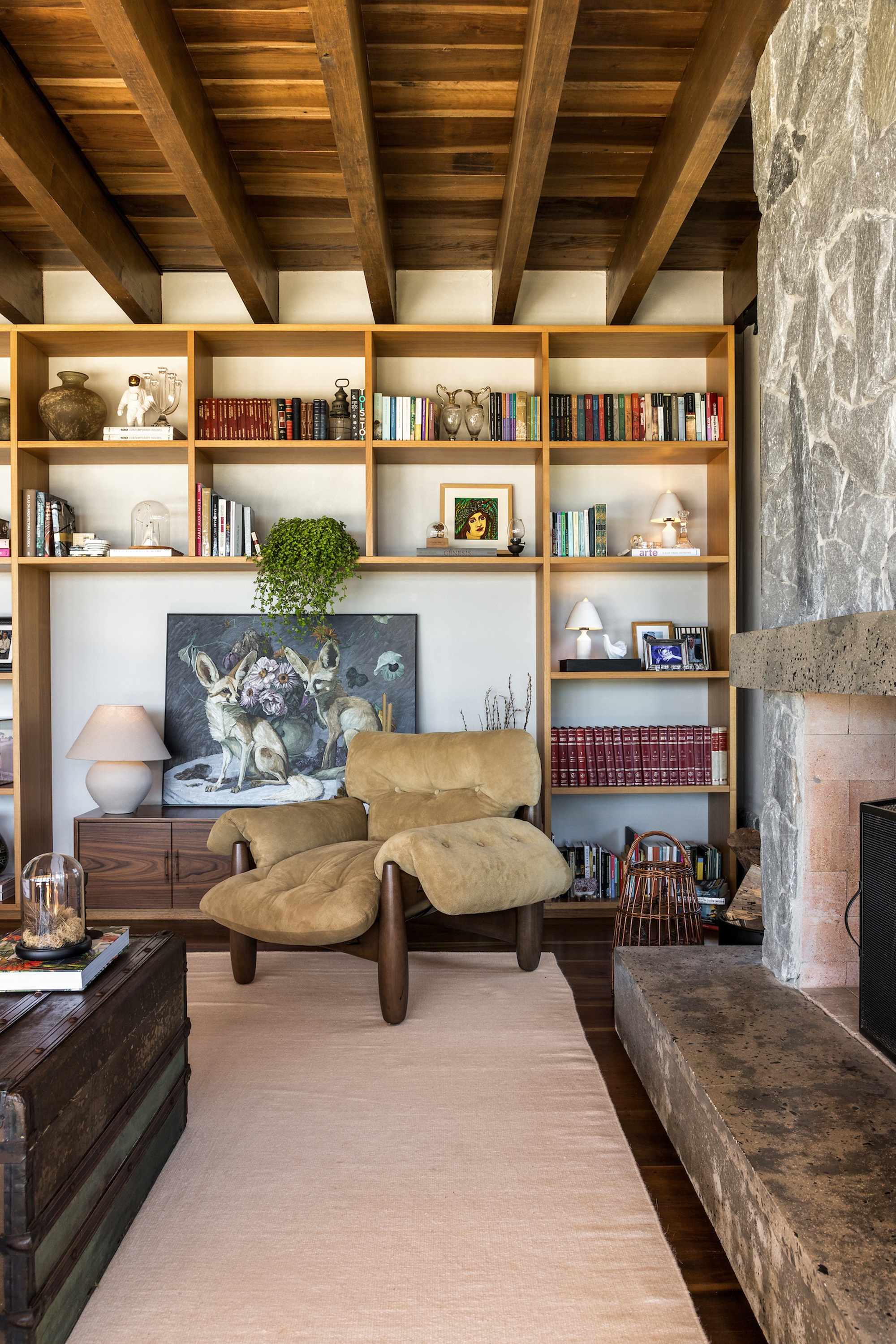 Timber-framed bookshelf wall with integrated lighting beside a stone fireplace and exposed ceiling beams