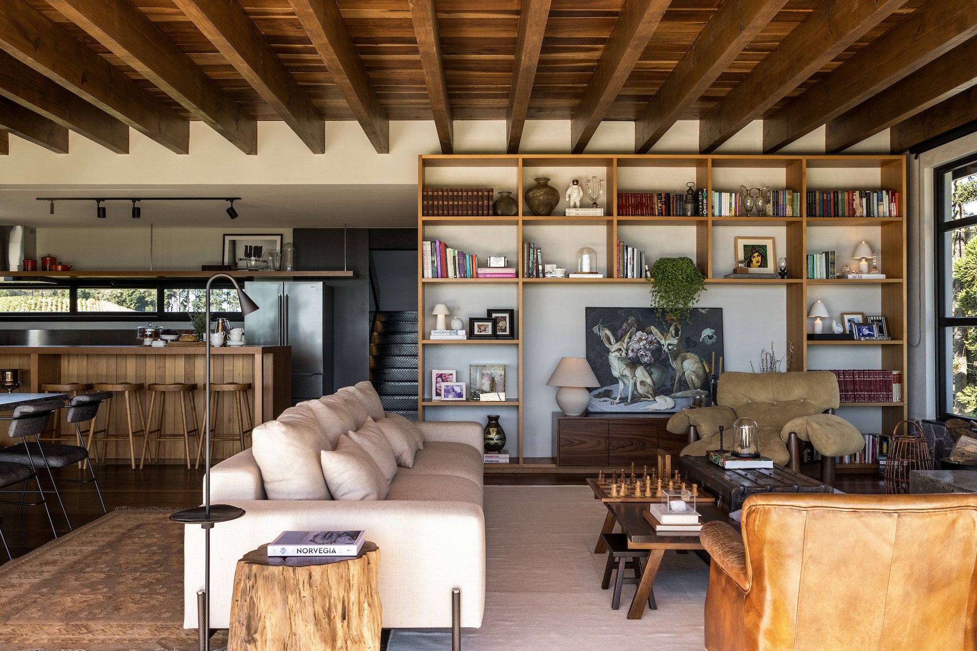 Open-plan living space with exposed timber structure linking built-in shelving, seating area, and kitchen beyond