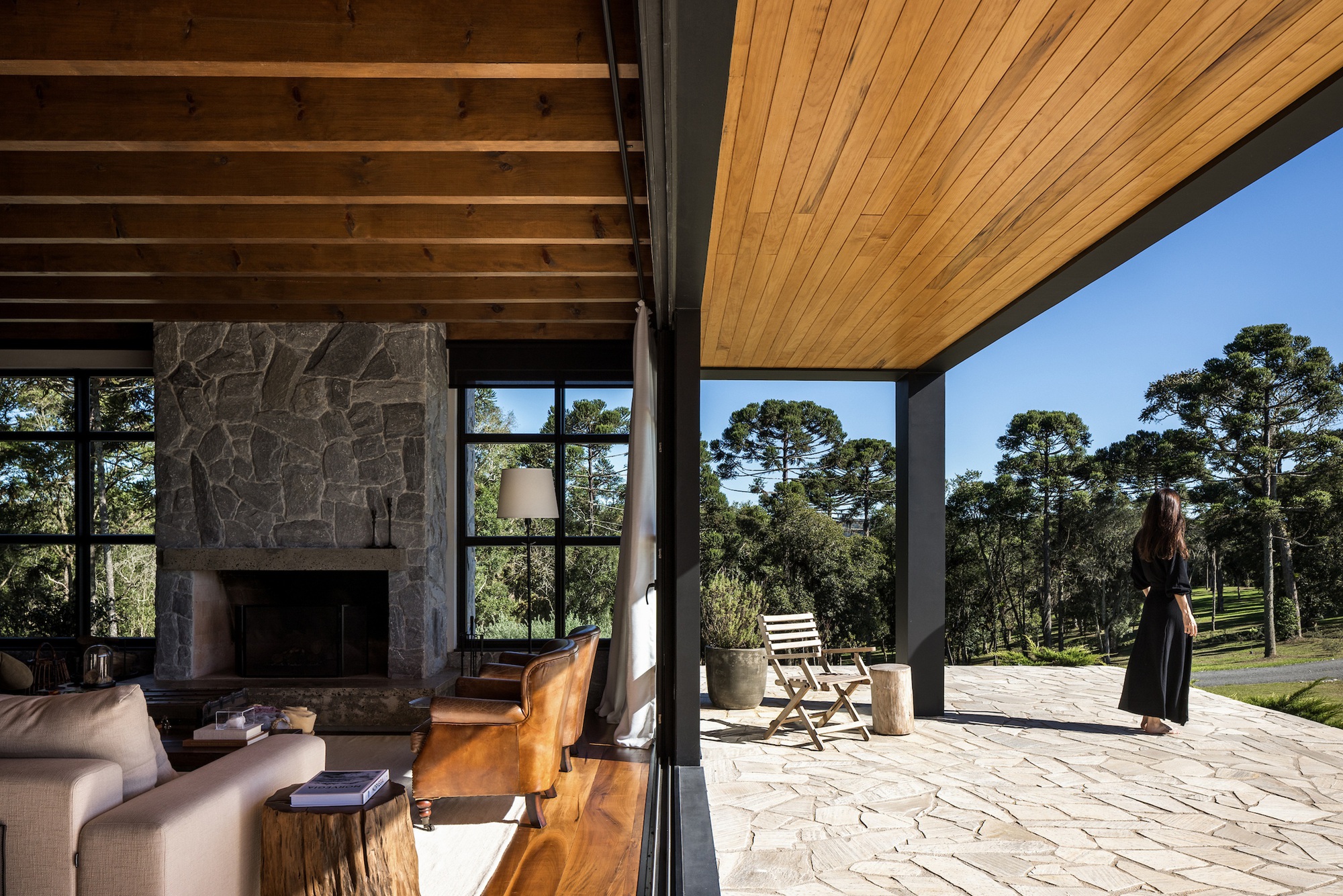 Covered terrace with stone fireplace opening to irregular flagstone paving and wooded landscape beyond