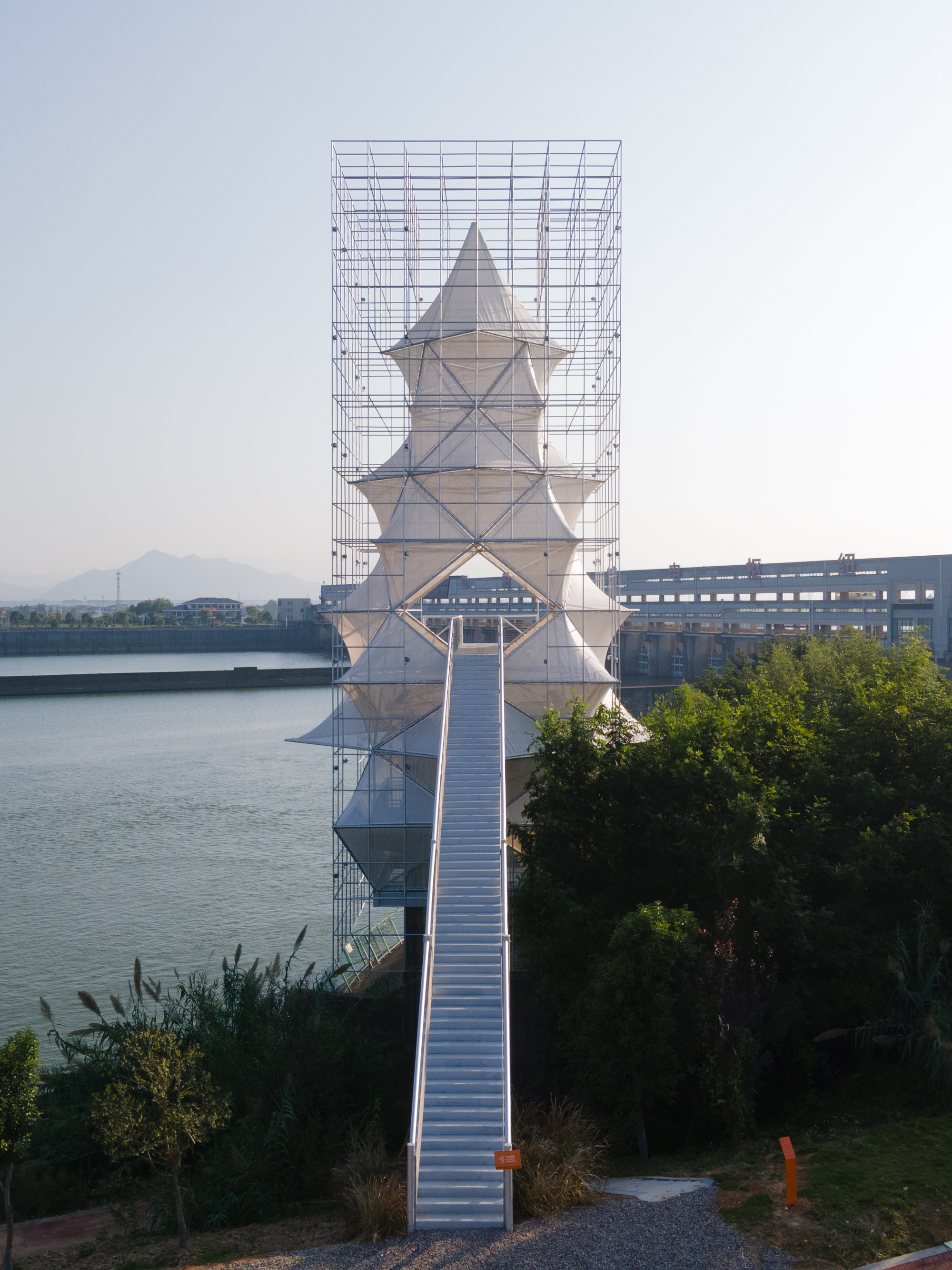 Tiered white tower enclosed by an outer steel scaffolding frame with a ramped pedestrian bridge at dusk