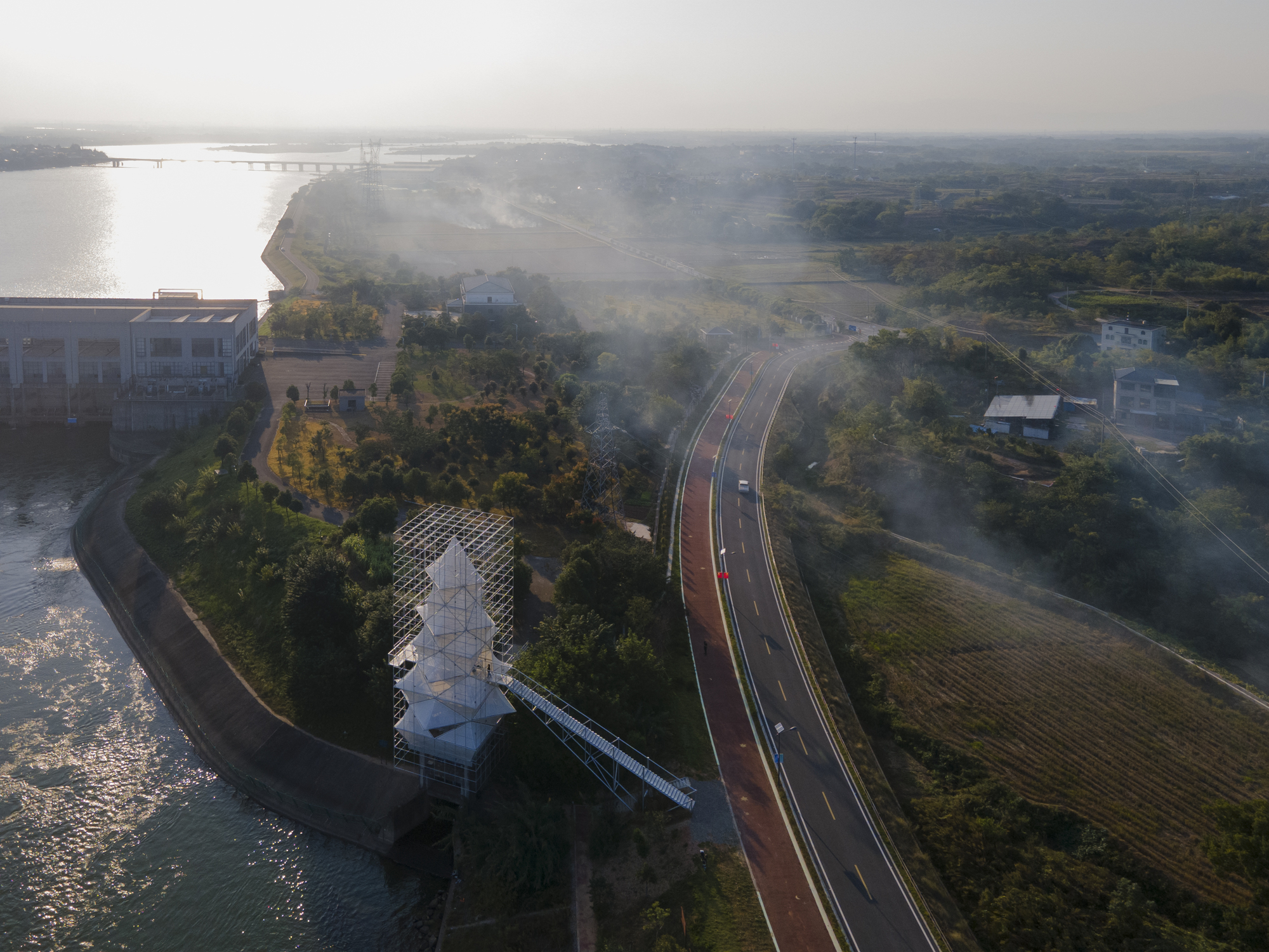 Aerial view of the scaffolded tower on a riverside peninsula beside a red bicycle path in morning mist