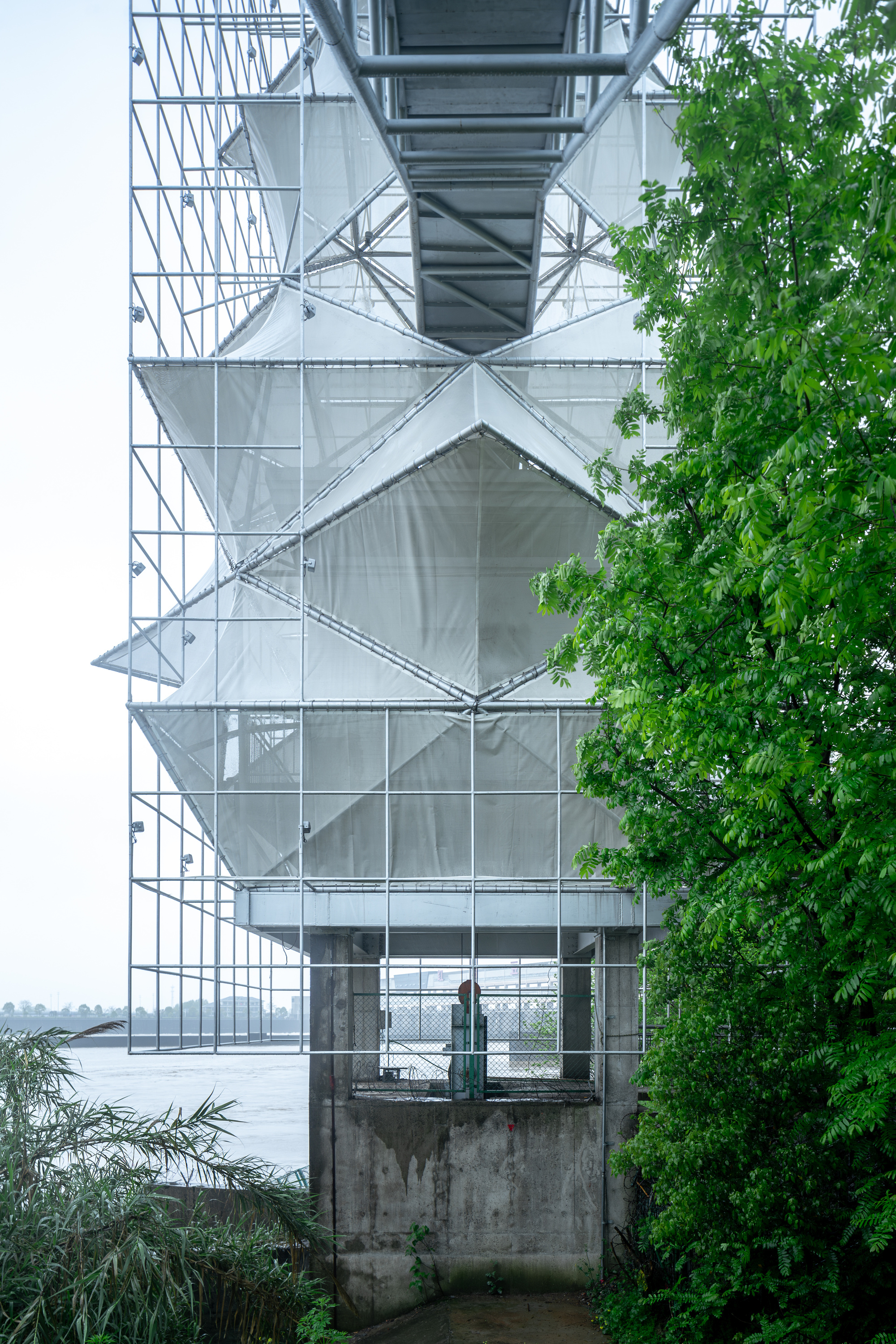 Base of the tower showing the concrete plinth and diagonal steel exoskeleton beside waterfront vegetation
