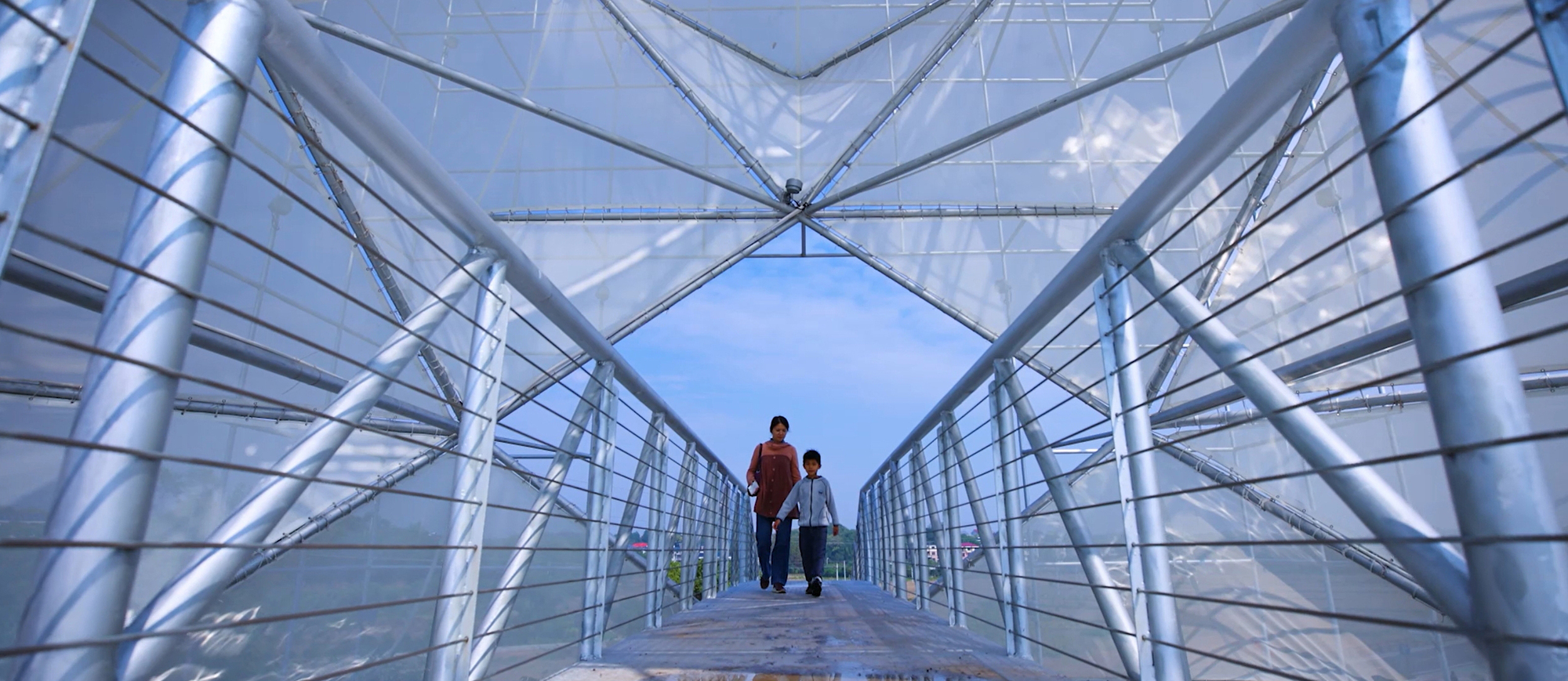 View along the ramped steel mesh bridge with two visitors walking toward the tower entrance at twilight