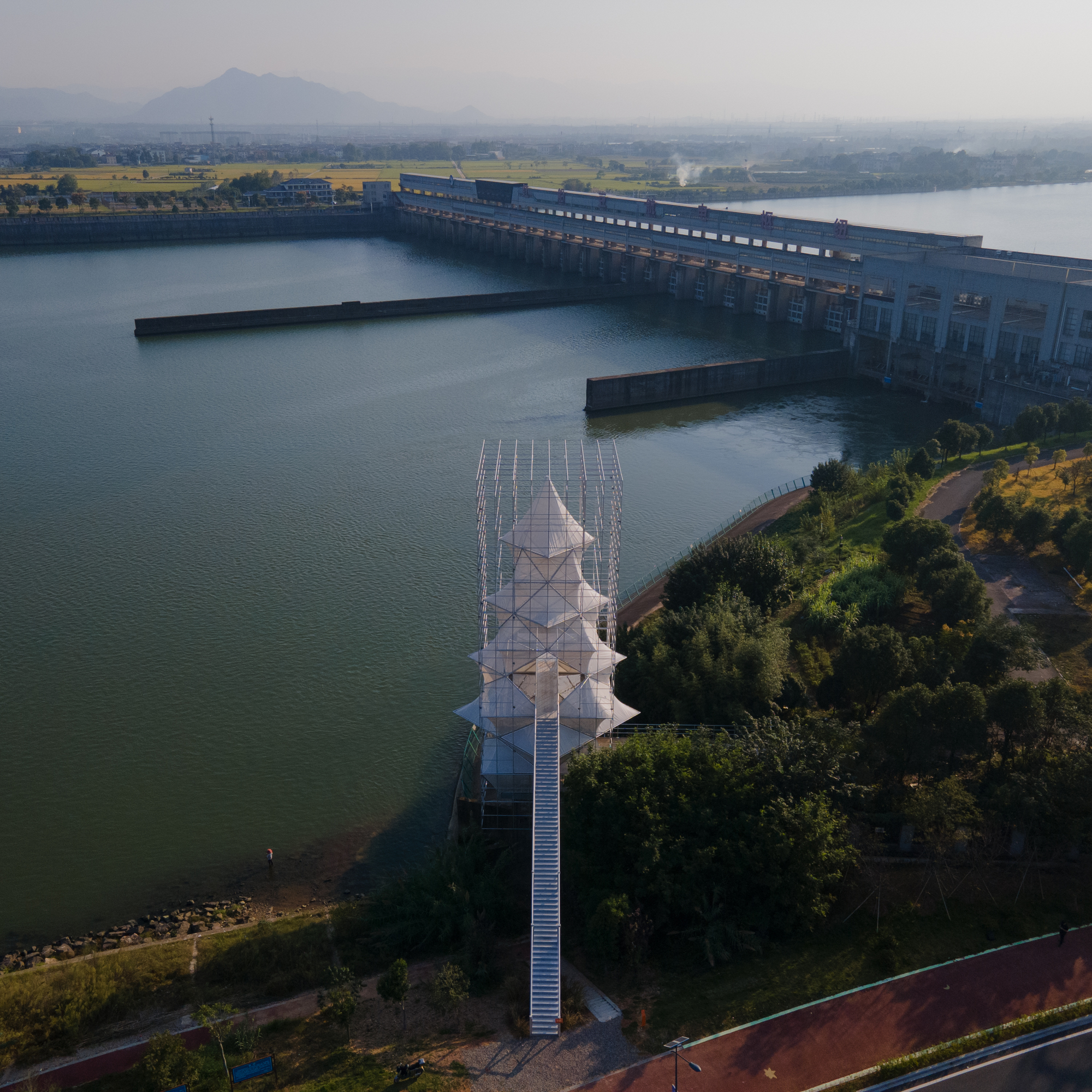 Aerial view of the white lattice tower and pedestrian bridge at the edge of the reservoir with dam in the distance