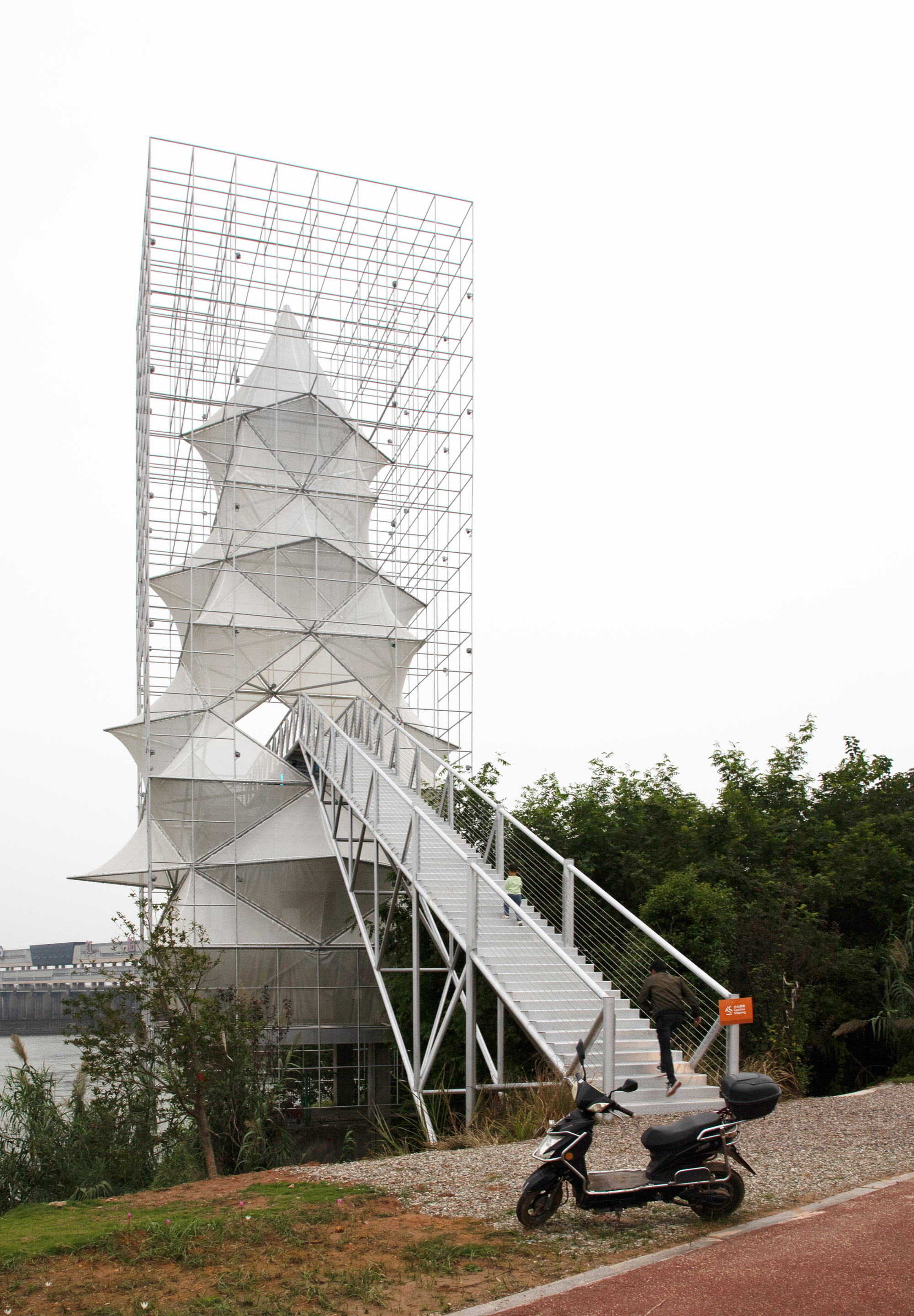 Side view of the steel scaffolding frame enclosing the stacked pyramidal volumes with a scooter parked below