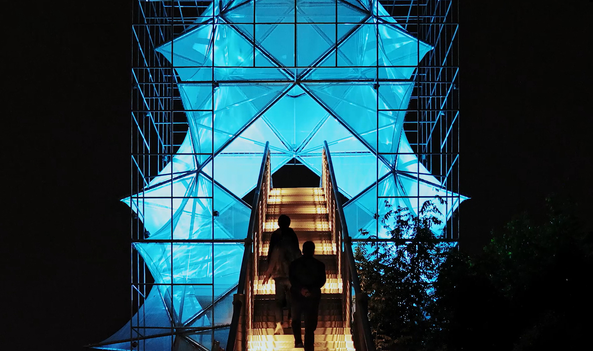 Nighttime view looking up through the illuminated blue glass panels and steel bracing with silhouetted figures on the stairs