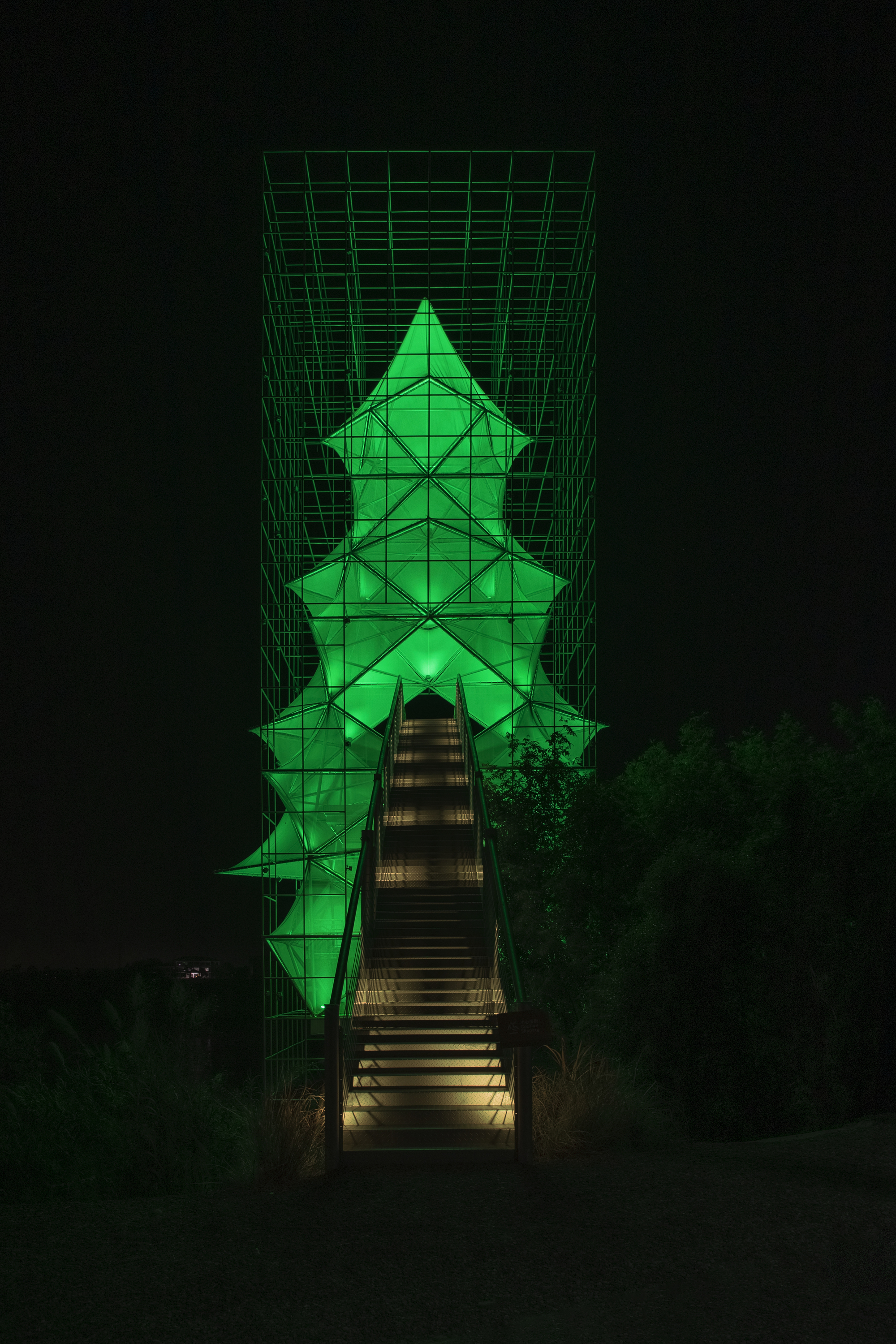 Illuminated pyramidal tower with green lighting and structural grid framing accessed by a central timber staircase at night