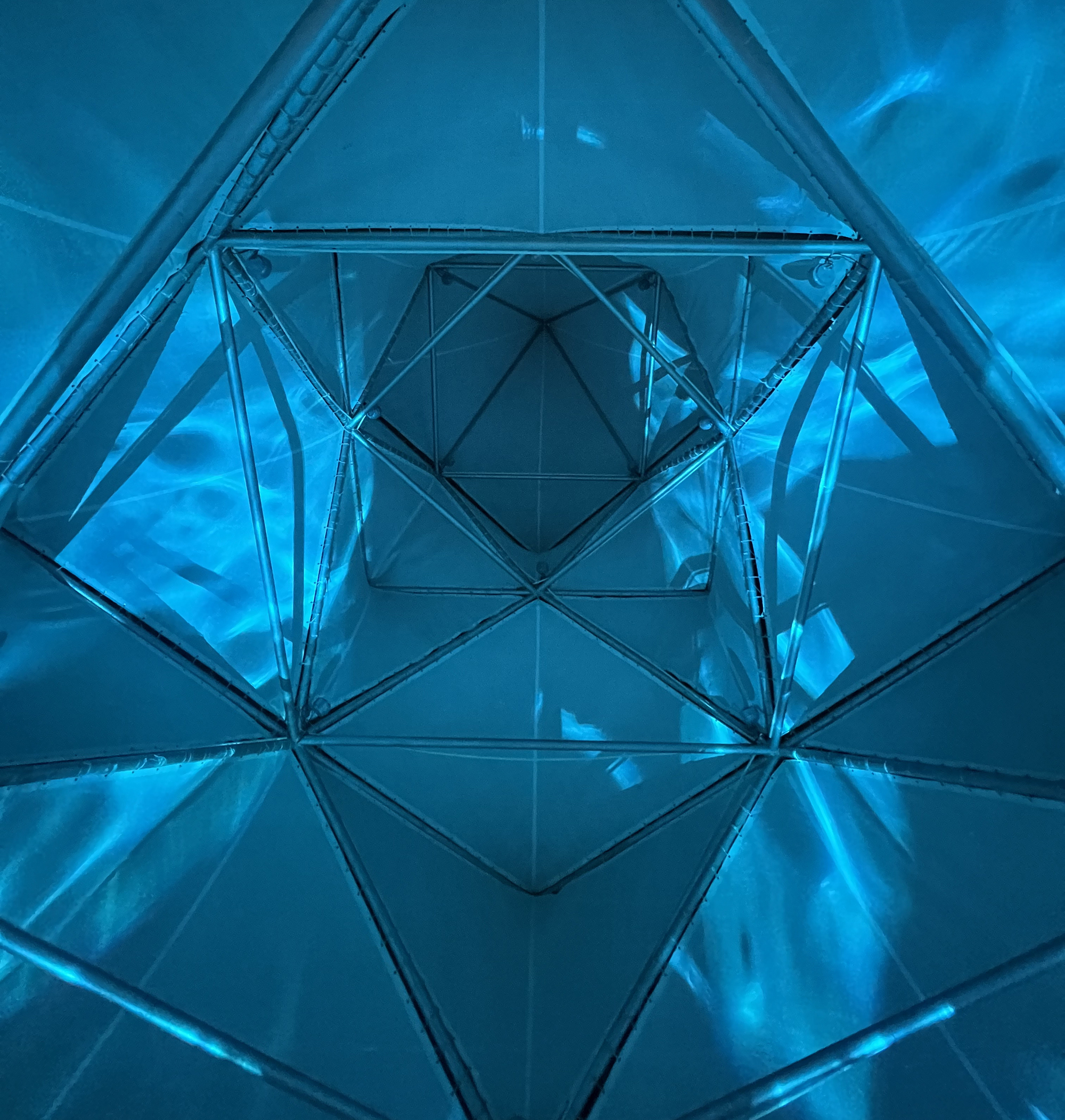 Interior view looking up through blue-lit triangulated glass and steel framework creating kaleidoscopic reflections