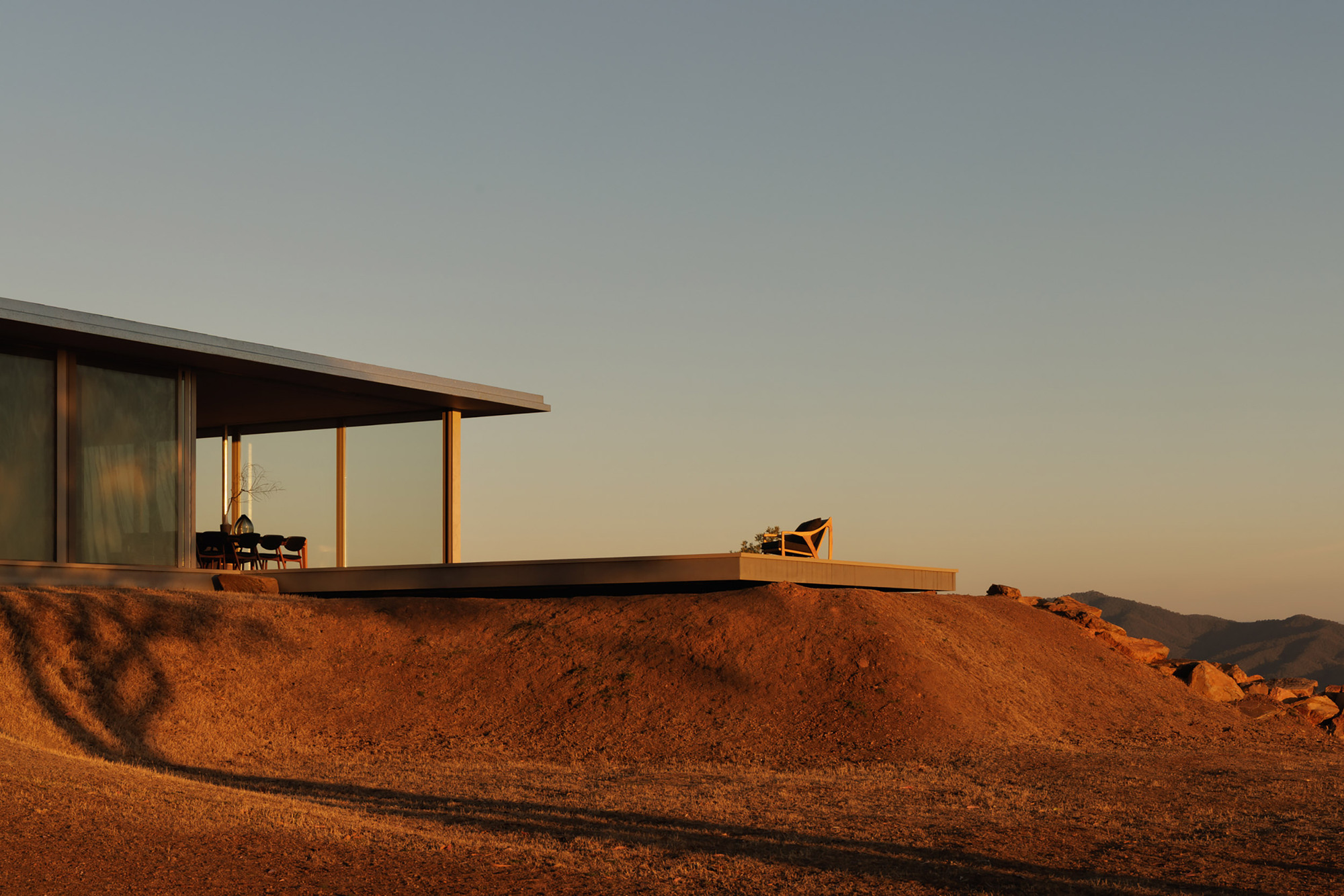Glass-walled pavilion emerging from the hillside with outdoor terrace and chair at golden hour