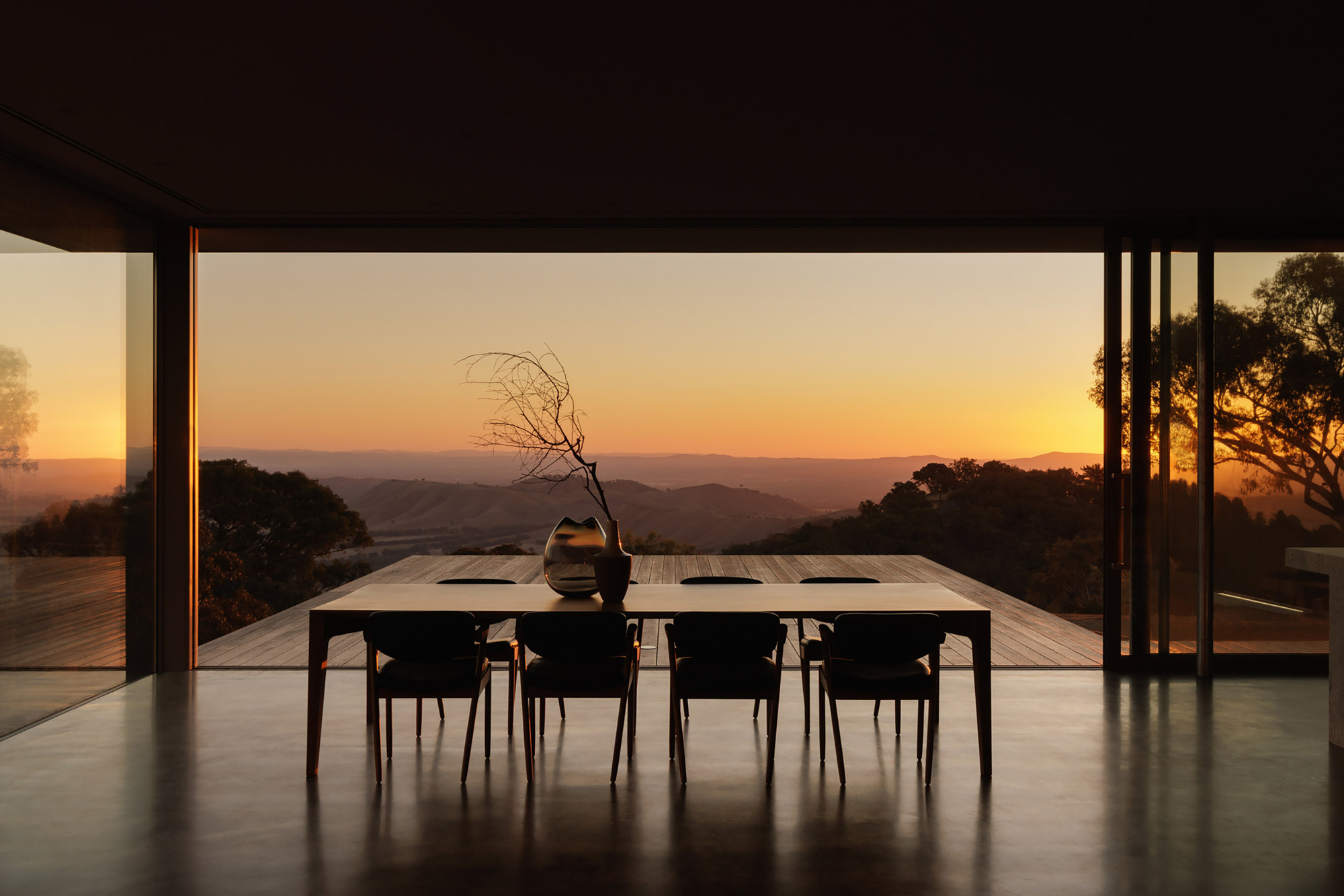 Dining table and chairs framed by full-height glass opening to deck and sunset vista