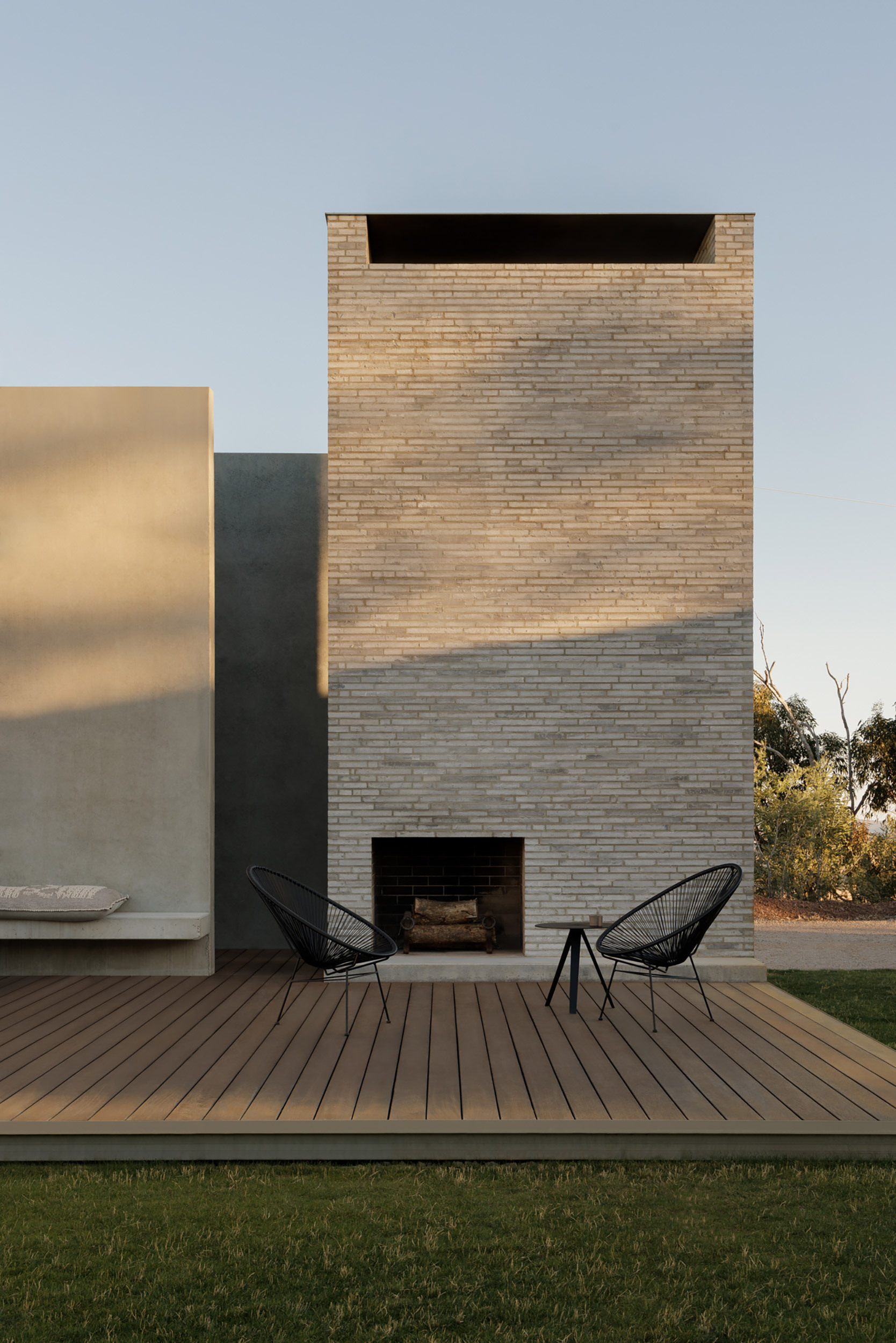 Two-story brick fireplace tower with horizontal opening framing a timber deck and pair of black wire chairs
