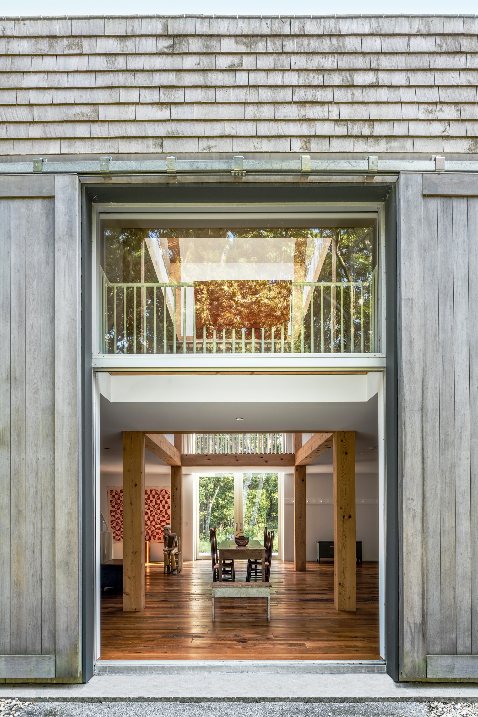 Exterior opening in weathered timber cladding revealing double-height interior with timber columns and balcony at dusk