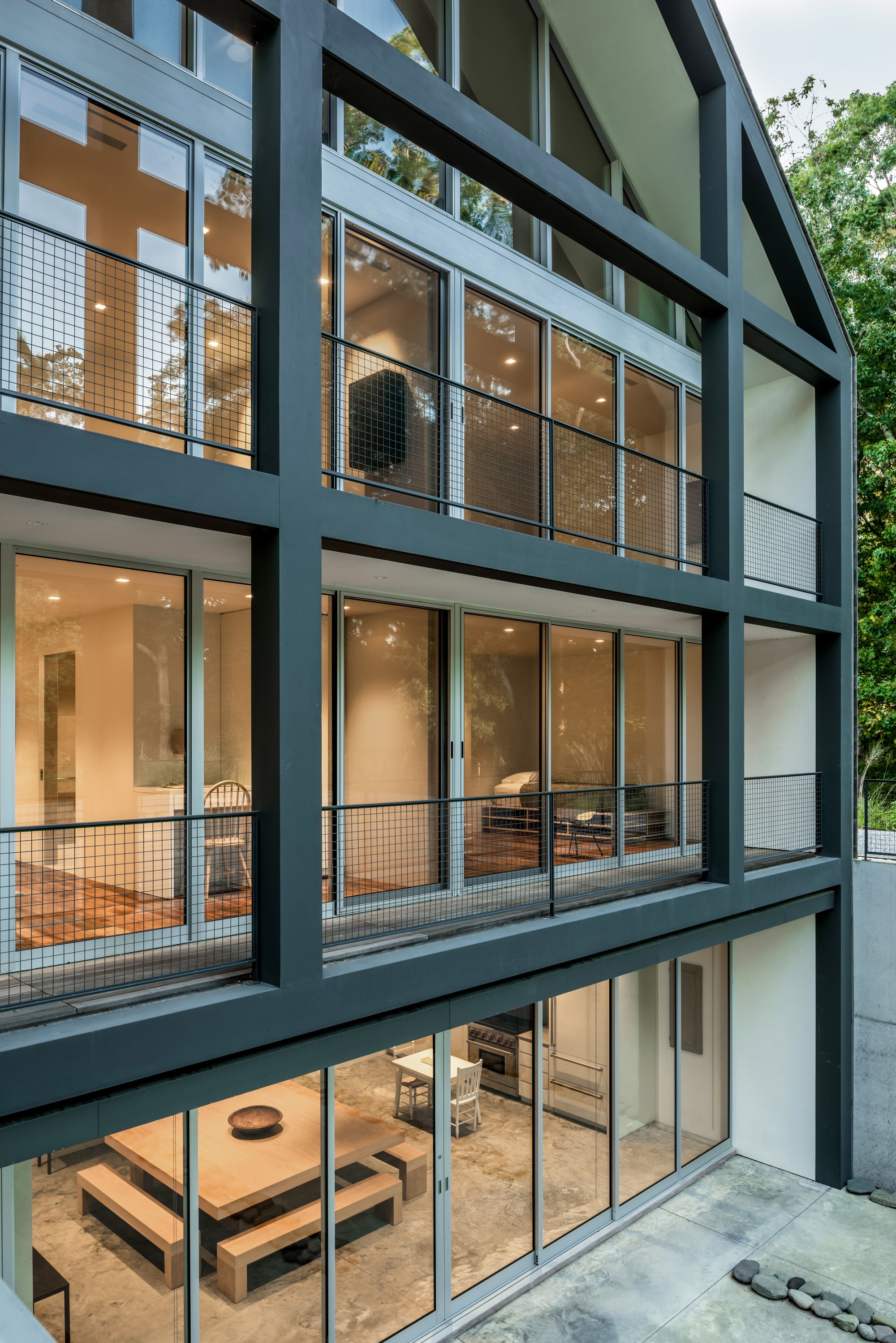 Multi-level facade with dark steel frames and wire mesh balcony railings at dusk beside leafy trees