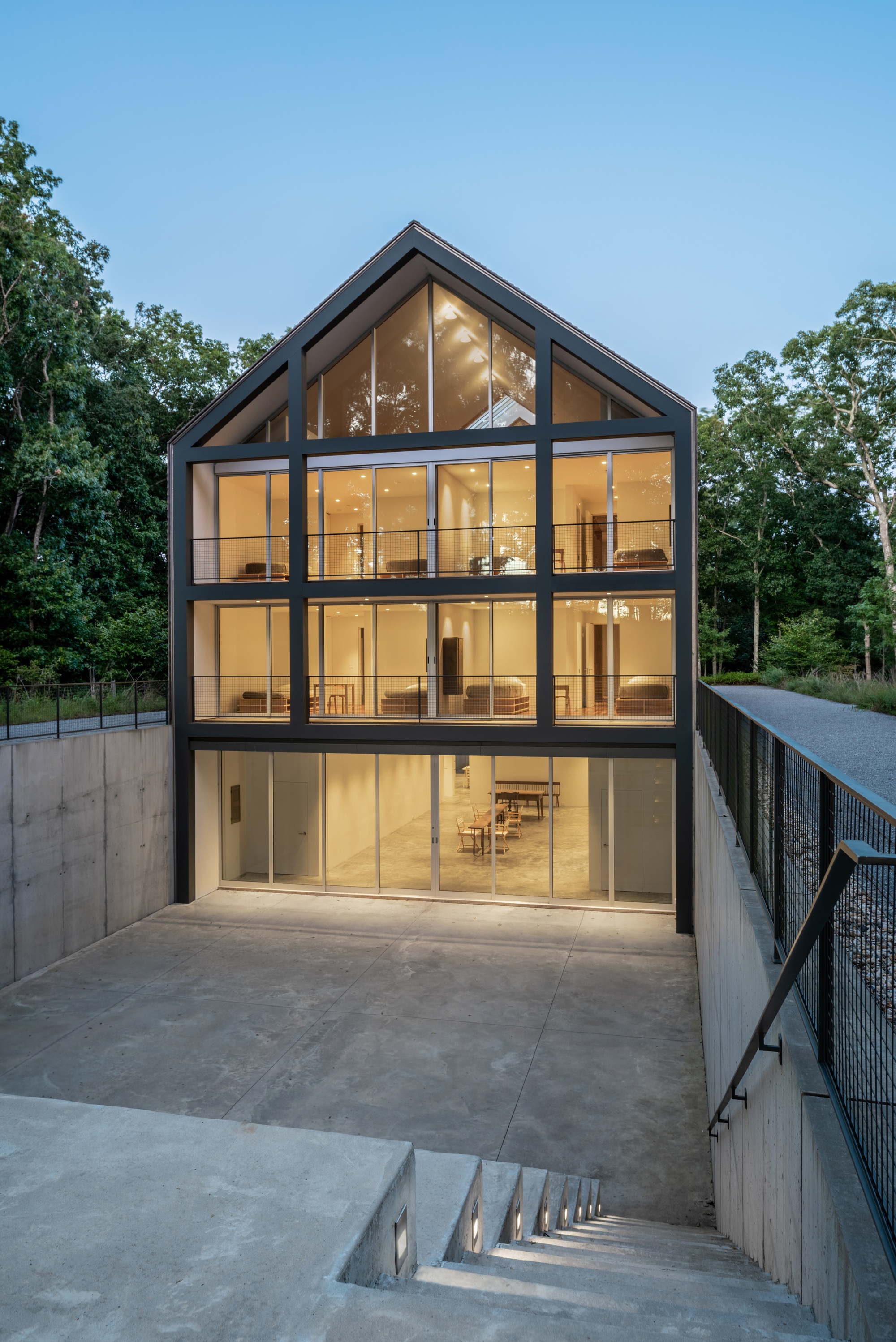 Gabled glass facade with black steel framing set into a concrete retaining wall at twilight