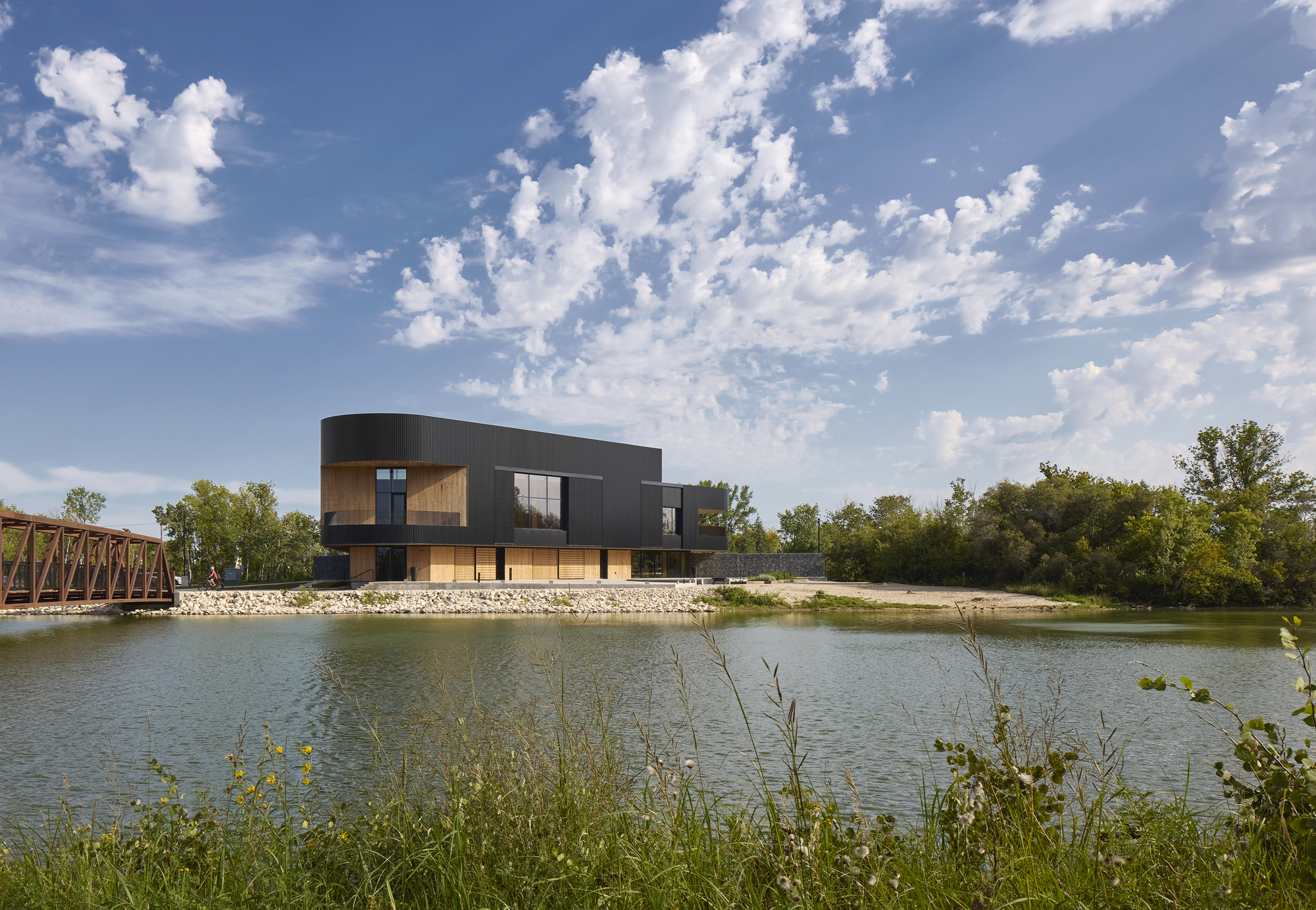 Two-story volume with curved metal cladding overlooking a pond with wildflowers and a truss bridge beyond