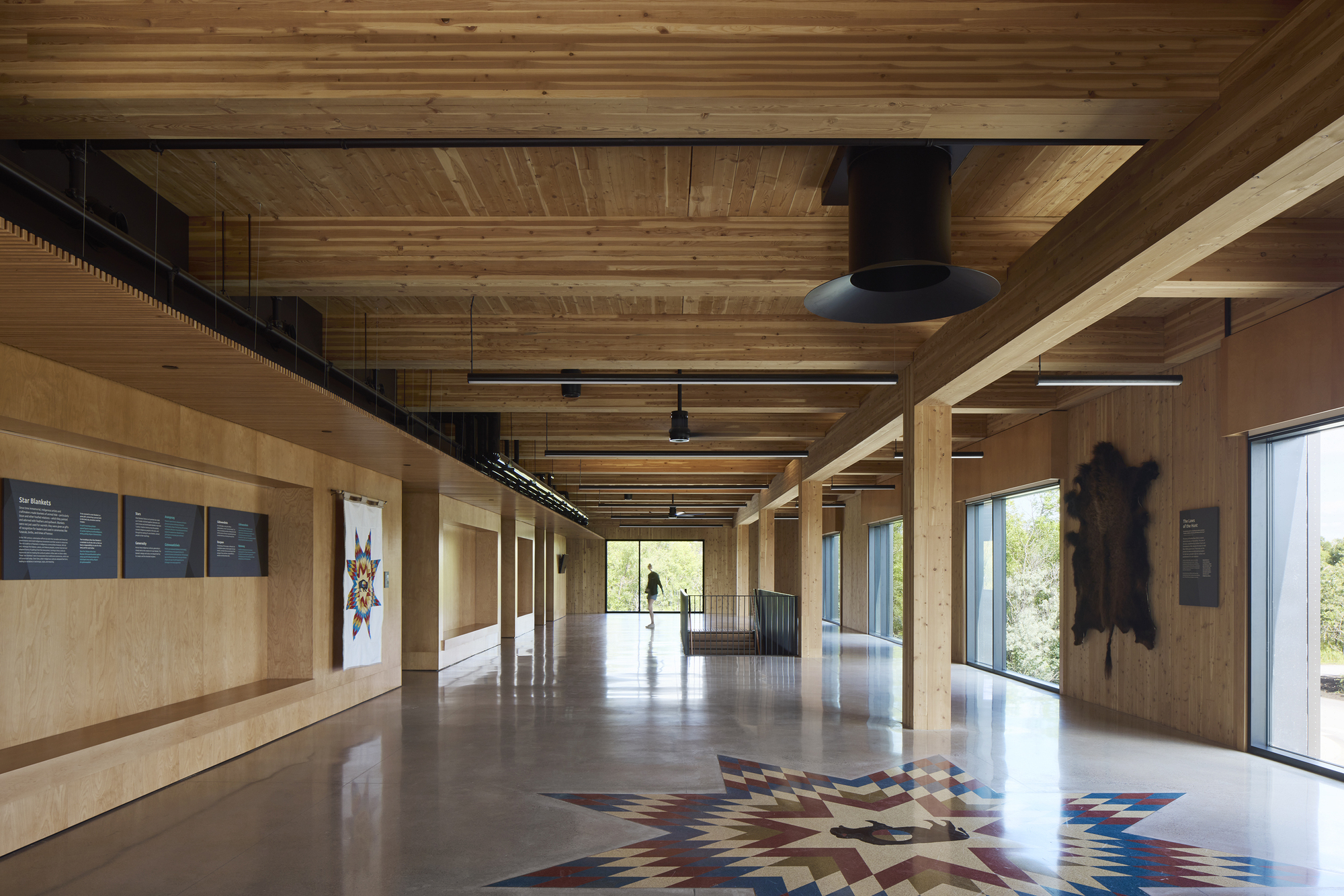 Interior gallery space with exposed timber beams and plywood walls displaying a patterned floor installation under linear lighting