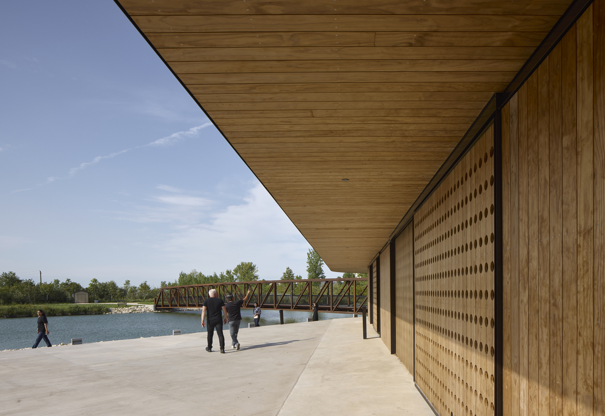 Covered porch with timber soffit and perforated wood screen wall overlooking the pond and steel truss bridge