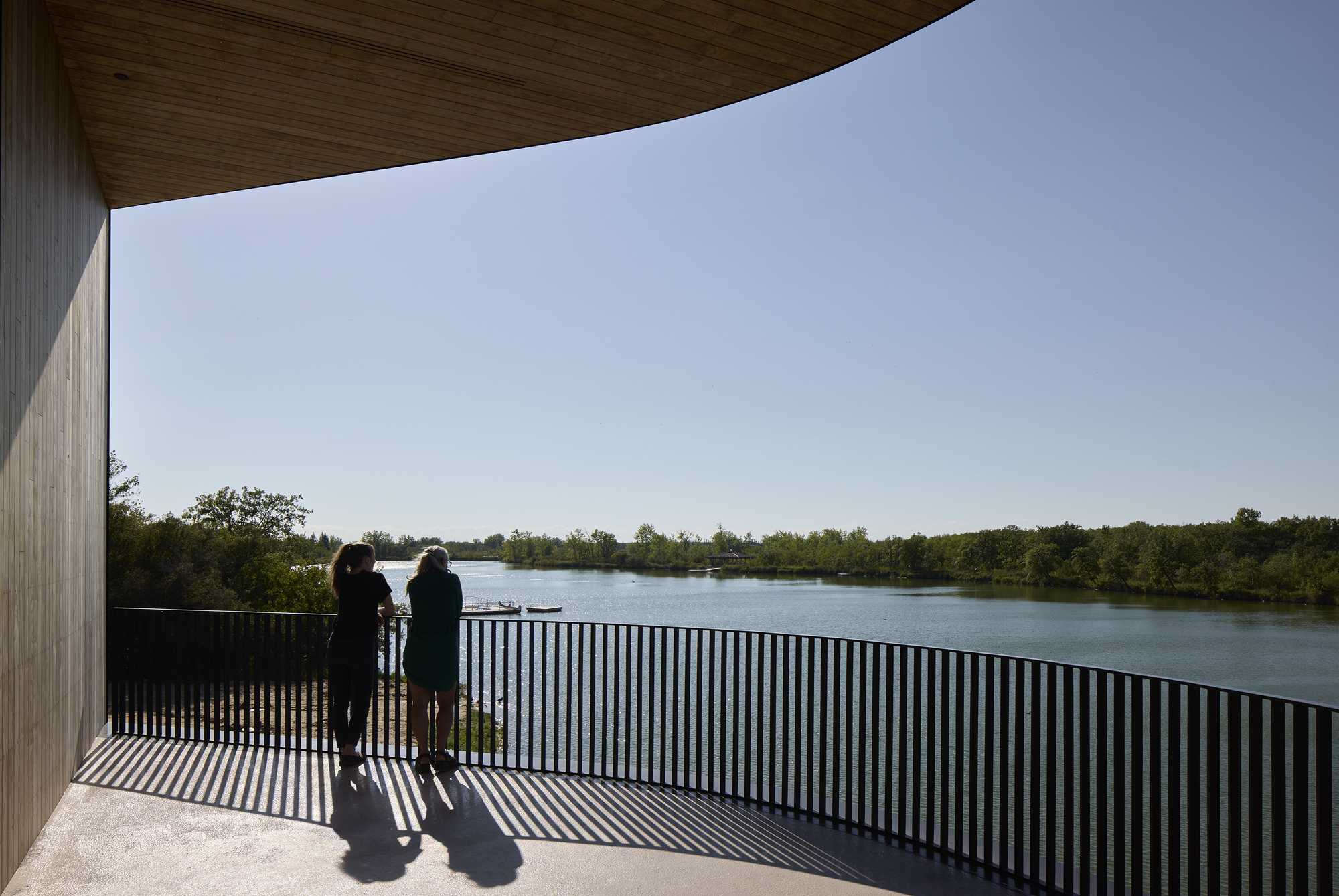 Covered terrace with vertical black railing and two visitors looking out toward wooded lakeshore