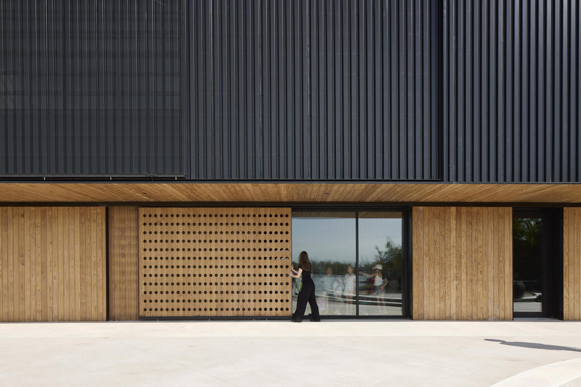 Ground-level facade with perforated timber panels and glazed openings beneath dark corrugated metal cladding as a person walks past