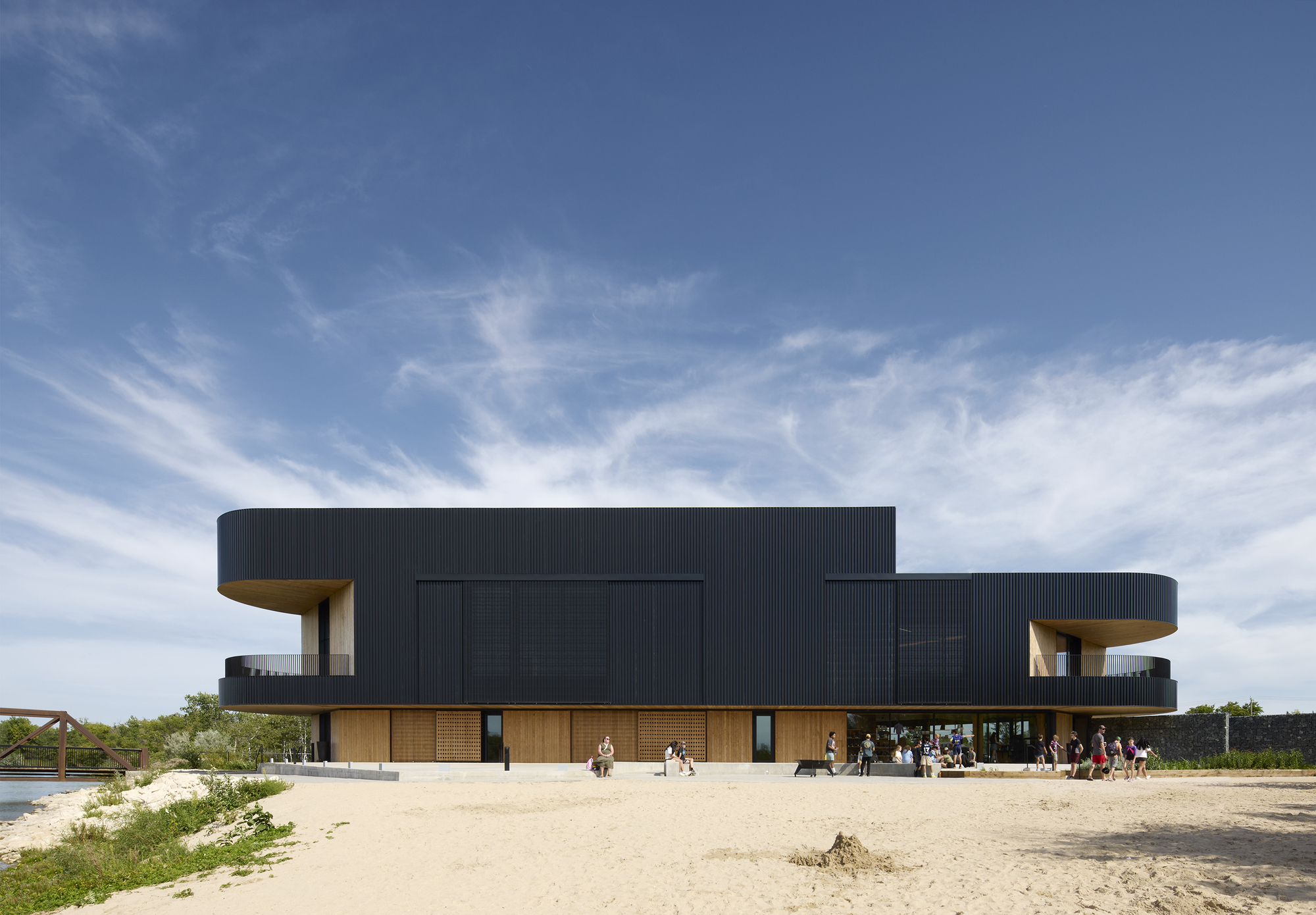 Two-story volume with black corrugated cladding cantilevering over timber base, set in sandy terrain with visitors gathering outside