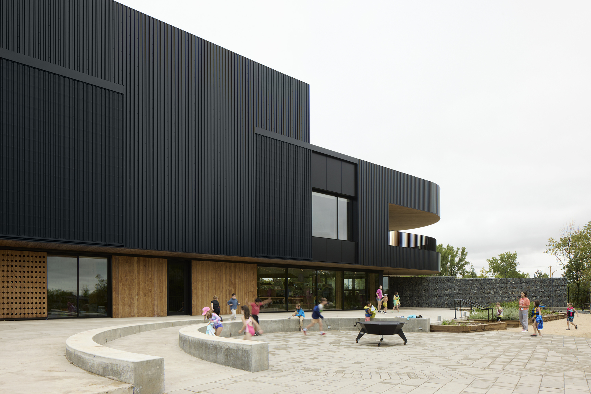 Curved corrugated facade with timber canopy overhangs and paved courtyard where children play around concrete benches