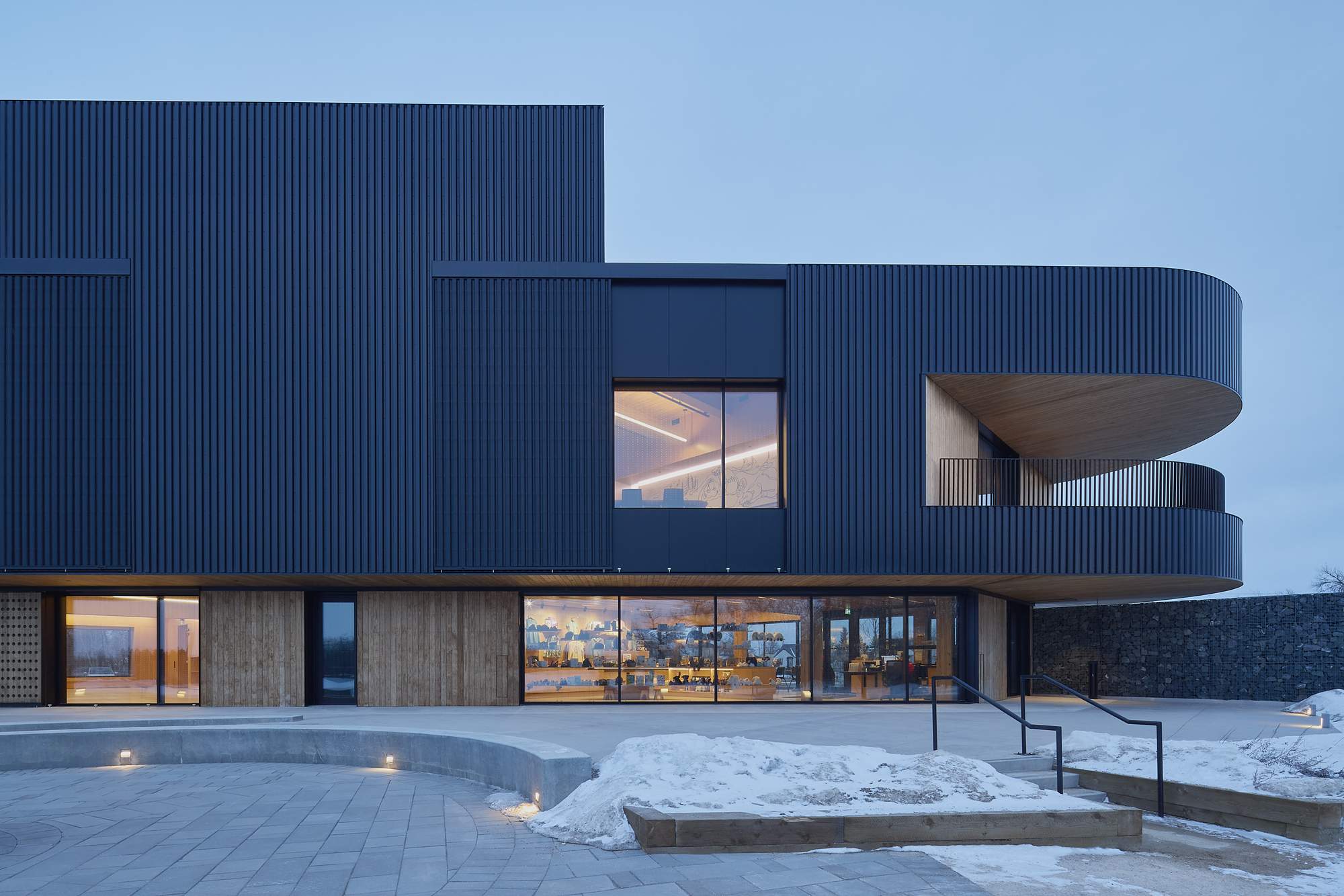 Ribbed blue metal facade with cantilevered upper volume illuminated at dusk in winter snow