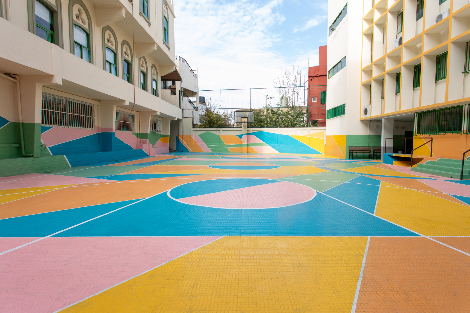 Basketball court with geometric pastel paving patterns viewed toward covered arcade and buildings beyond